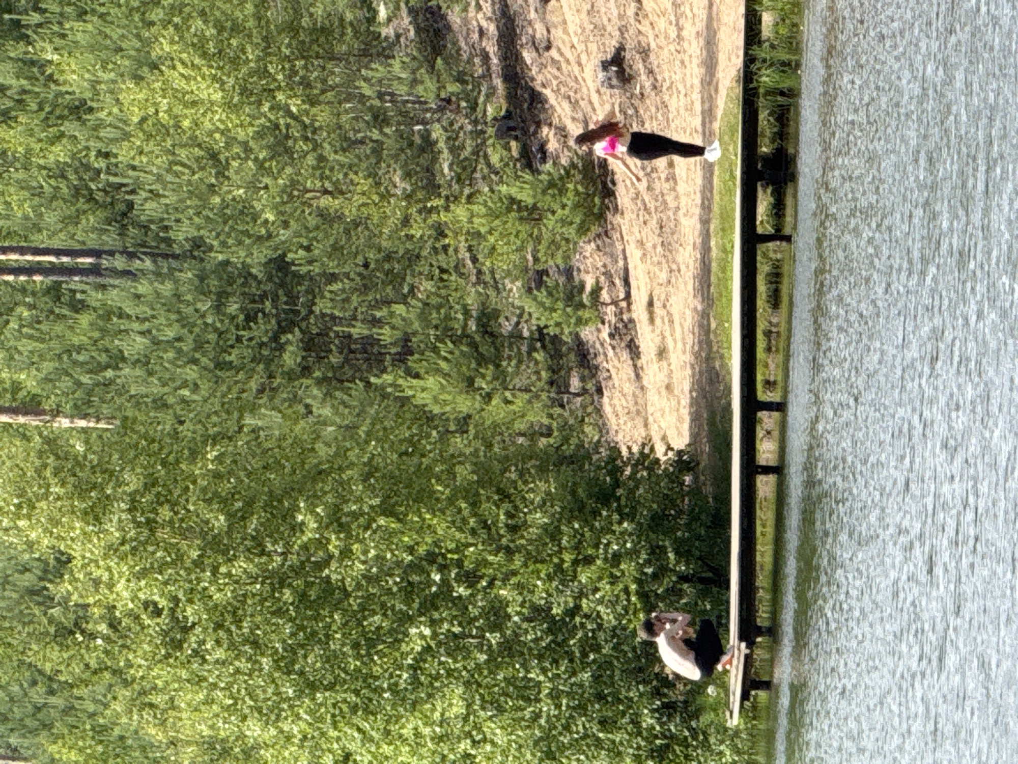 Two individuals are present on a wooden pier extending over a body of water. A person is crouched on the left side of the pier, holding a camera, seemingly taking a photograph. On the right side of the pier, a person stands with their arms outstretched, facing away from the camera. The background consists of a wooded area with numerous pine trees on a sandy slope. The location is identified as the city of Peski, Russia.