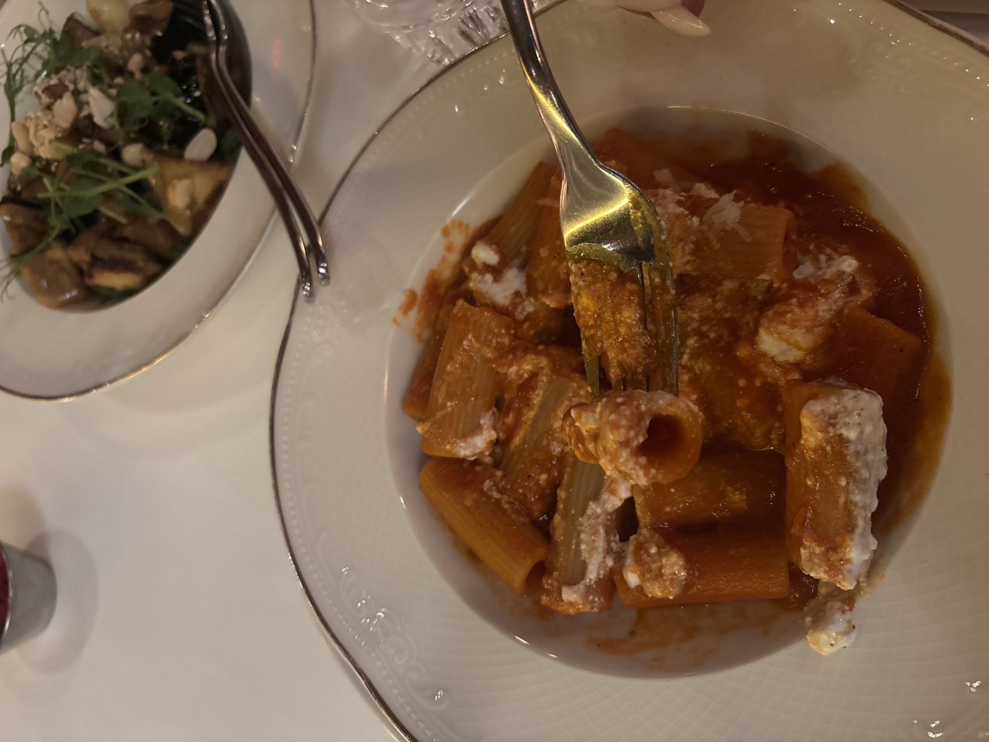 The image shows two plates of food on a white table. The plate on the right contains pasta in tomato sauce, with ricotta cheese. A fork pierces a piece of pasta and lifts it from the plate. The plate on the left features a salad with greens, mushrooms, and nuts. A fork and spoon rest on the edge of the salad plate. The scene appears to be indoors, likely in a restaurant. There is no immediate evidence to confirm the location is Yerevan, Armenia.