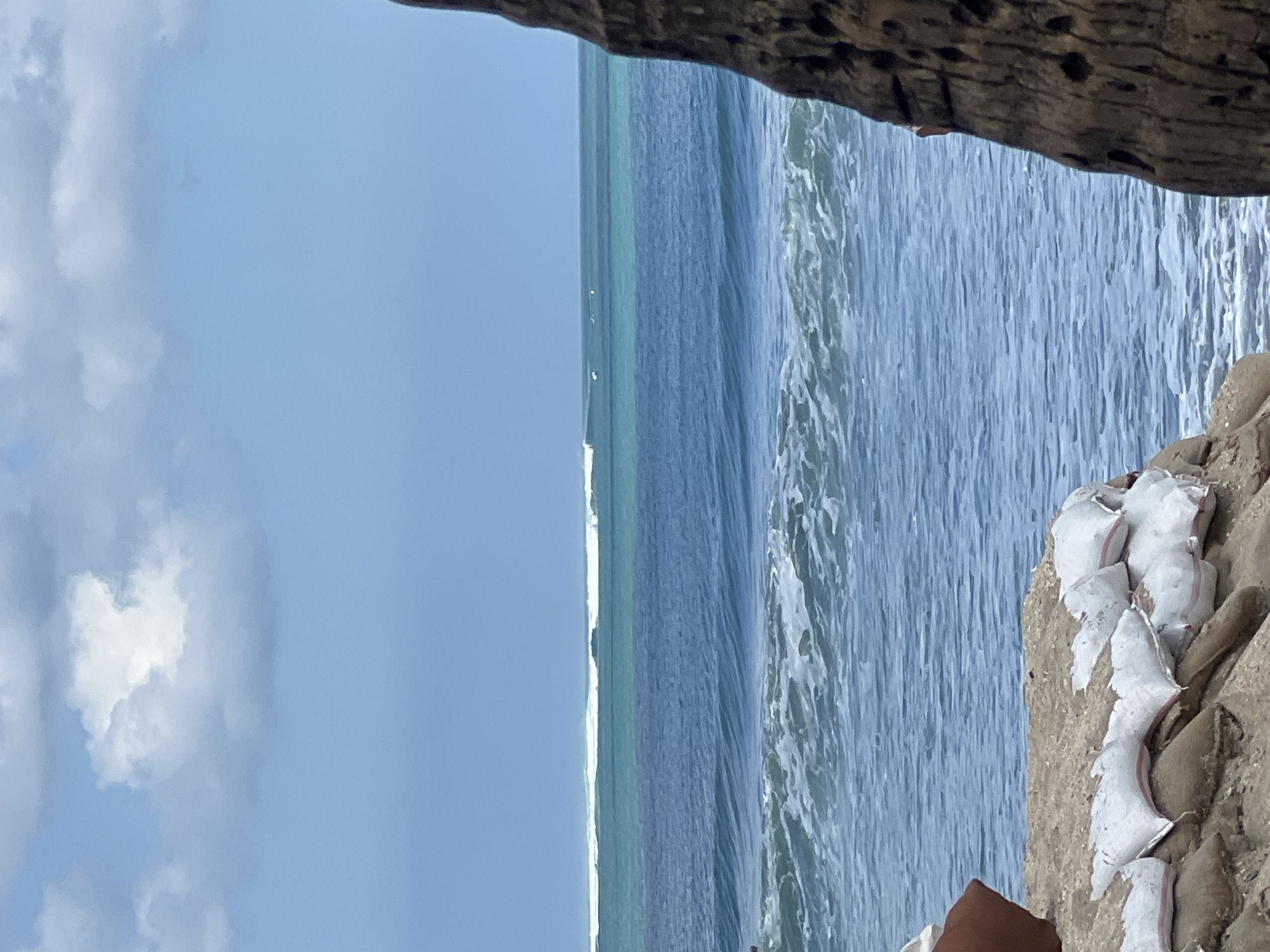 The image depicts a coastal scene in Kuta, Bali, Indonesia. The foreground shows sandbags placed on a rocky shore, suggesting coastal protection measures. In the midground, the ocean exhibits multiple sets of waves. The furthest set of waves appears to be breaking, with several surfers visible in the distance. The water transitions from a deep blue further out to a lighter turquoise closer to the shore. The sky above is a clear blue, with a few scattered white clouds. A portion of a textured, dark brown tree trunk is visible on the right side of the frame, adding to the tropical setting.