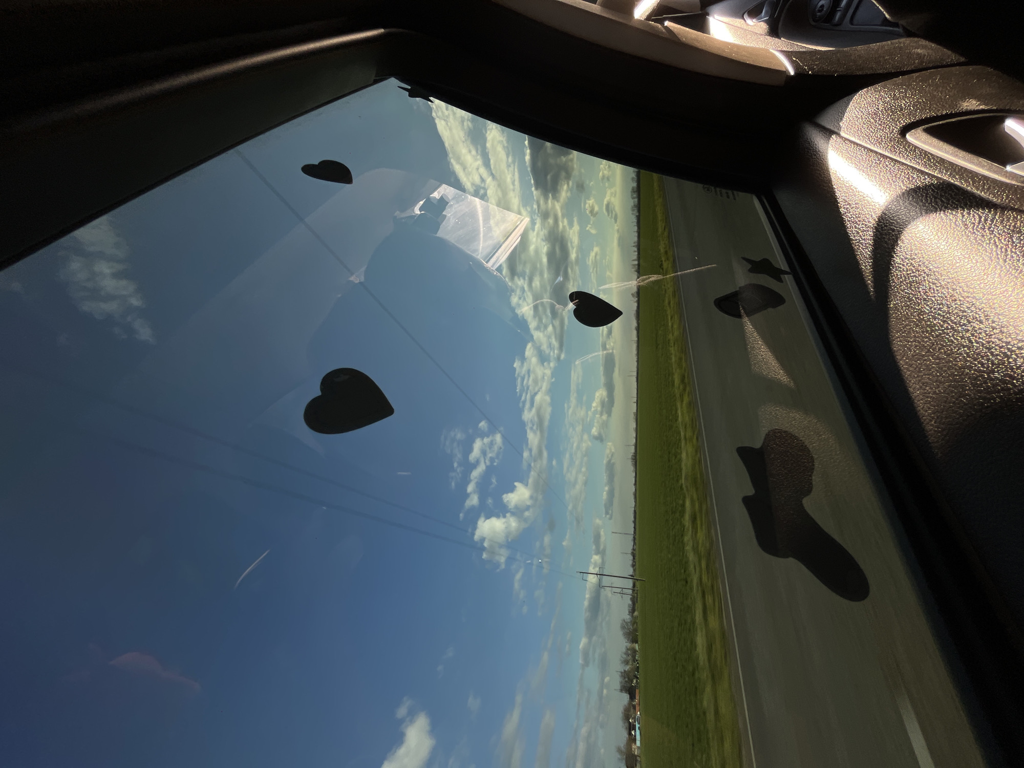 The image captures a bright, sunny outdoor scene viewed through the side window of a vehicle, rotated 90 degrees clockwise. The perspective is from inside the car, looking out at a rural or semi-rural landscape.
In the foreground, the dark, textured plastic interior of the car's door panel is visible on the right, illuminated by strong sunlight that creates distinct highlights. The top edge of the window frame is also visible at the top.
The window itself is adorned with several dark, silhouetted shapes, likely stickers or suction cups. These include at least three heart shapes of varying sizes, a star shape, and a footprint shape, suggesting a personalized or child-friendly decoration. Reflections of clouds and possibly a rectangular object from inside the car are also visible on the glass.
Beyond the window, a vast expanse of clear blue sky with scattered white cumulus clouds dominates the upper half of the view. Below the horizon, a flat, green field stretches into the distance, bordered by what appears to be a paved road or shoulder on the right. Several utility poles with overhead lines traverse the landscape, indicating infrastructure in the area. In the far distance, faint outlines of trees or structures are visible.
The time of day appears to be midday or afternoon due to the bright, direct sunlight. The weather is clear and sunny. While specific location cues are limited to a general rural or highway setting, the context provided indicates this is in Ternovskaya, Russia. There is no visible text in the image. The decorations on the window add a touch of warmth or playfulness to the travel scene.