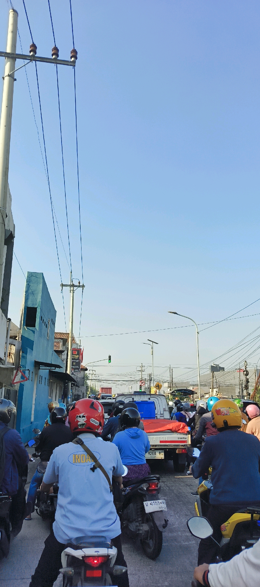 The image captures a traffic jam in Gedangan, Indonesia, viewed from the perspective of someone riding a motorcycle. A line of motorcycles and cars is stopped on a street lined with buildings and power lines. The sky is clear and blue. Several motorcyclists are visible, many wearing helmets in various colors, including red, blue, yellow, and black. One person in the foreground wears a light blue t-shirt with the word "INDAT" visible on the back. A red truck with a blue container and a red tarp covers is in the middle of the traffic. Traffic lights are visible in the distance, indicating a controlled intersection. The overall scene depicts a typical urban traffic situation.