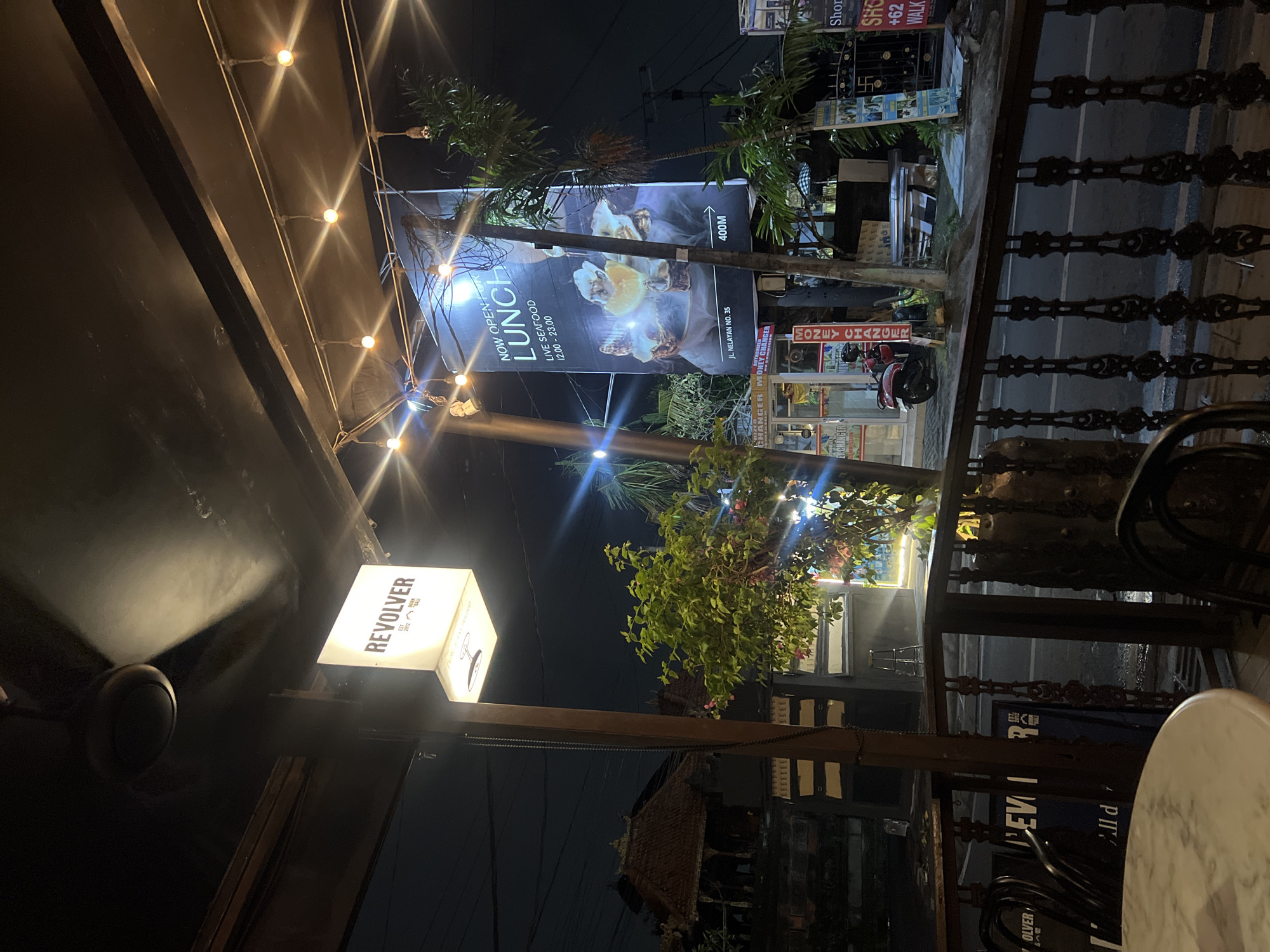 In North Kuta, Indonesia, at night, a view from an outdoor seating area looks across a street. A white sign illuminated from within, bearing the text "REVOLVER EST 2012 BALI" in black, hangs from a pole to the left. Above, a dark ceiling is visible with a ceiling fan. String lights with exposed bulbs are hung across the top of the area. A large billboard displays an advertisement for "NOW OPEN FOR LUNCH LIVE SEAFOOD 12.00 - 23.00" with an image of oysters and lemons. Below the billboard, signs for "MONEY CHANGER" are visible. A red scooter is parked on the street. The area is bordered by a wooden railing. In the foreground, a round marble-topped table is partially visible with dark chairs. The street and surrounding buildings are dimly lit by ambient light and streetlights. Trees and foliage are also present in the scene.
