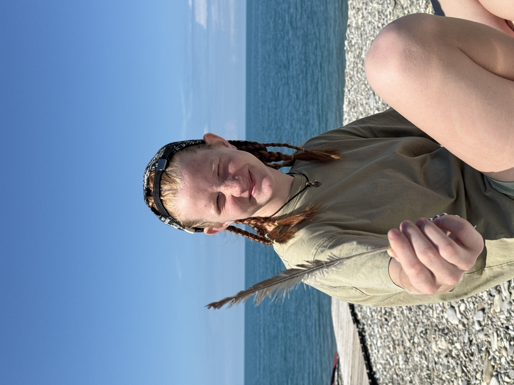 A young person with freckles and auburn hair styled in braids sits on a pebble beach, holding a feather in their outstretched hand. They are wearing a light green t-shirt, shorts, and a patterned bandana on their head. The background features the calm, blue sea and a clear sky. A wooden structure is visible on the left side of the image. The location is identified as Nebug, Russia.