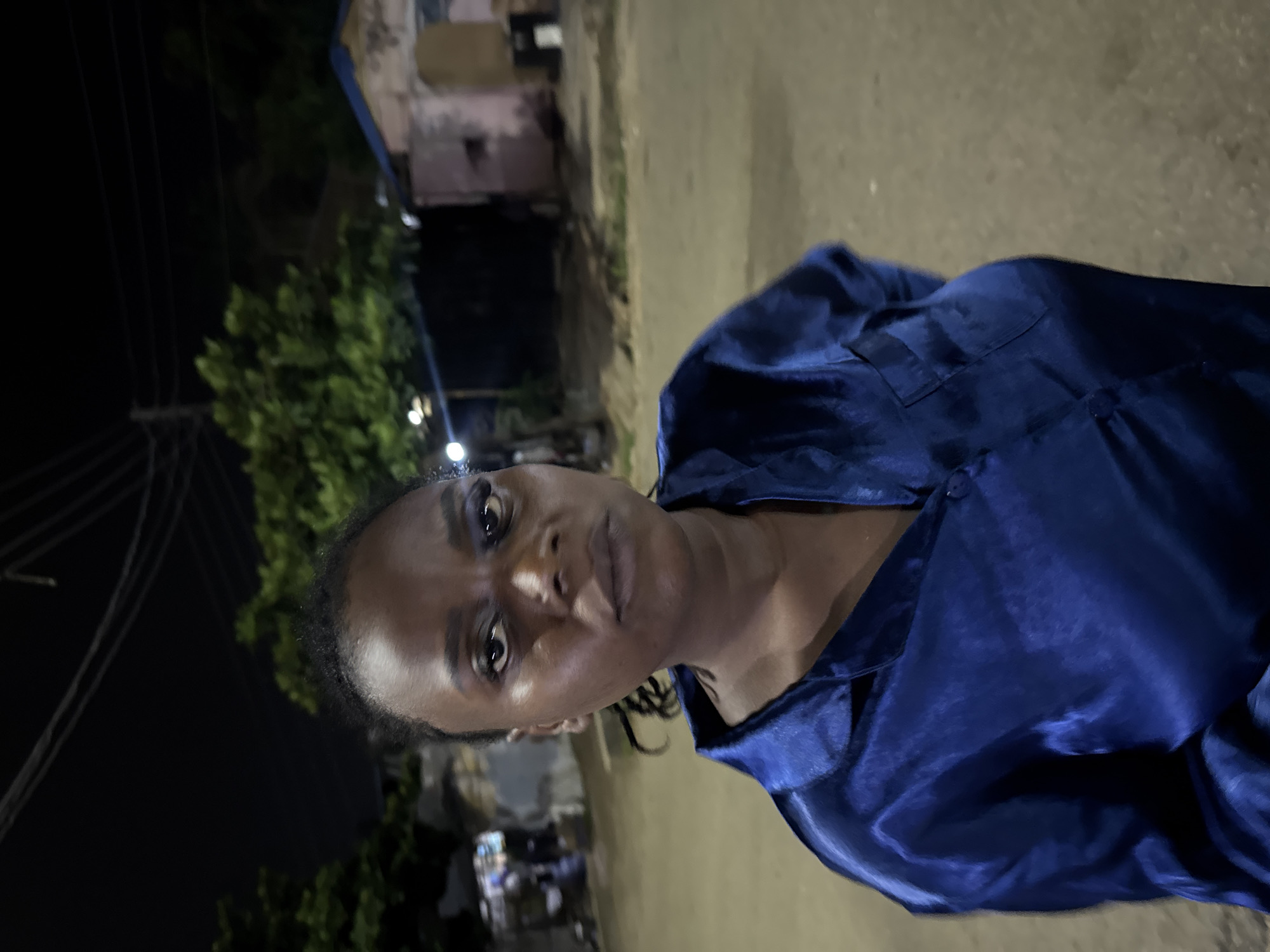 A medium shot captures a Black woman with braided hair and dark skin standing outdoors at night in Abuja, Nigeria. She is looking directly at the camera with a neutral expression. She is wearing a shiny, dark blue, long-sleeved collared shirt, possibly silk or satin, buttoned at the front. The background features a dirt road, some trees, and a building with a pinkish-tan facade and a light blue roof. Overhead power lines are visible against the dark sky. The lighting suggests artificial illumination, likely from streetlights, casting a warm glow on the scene. The environment appears to be a residential or semi-urban area.