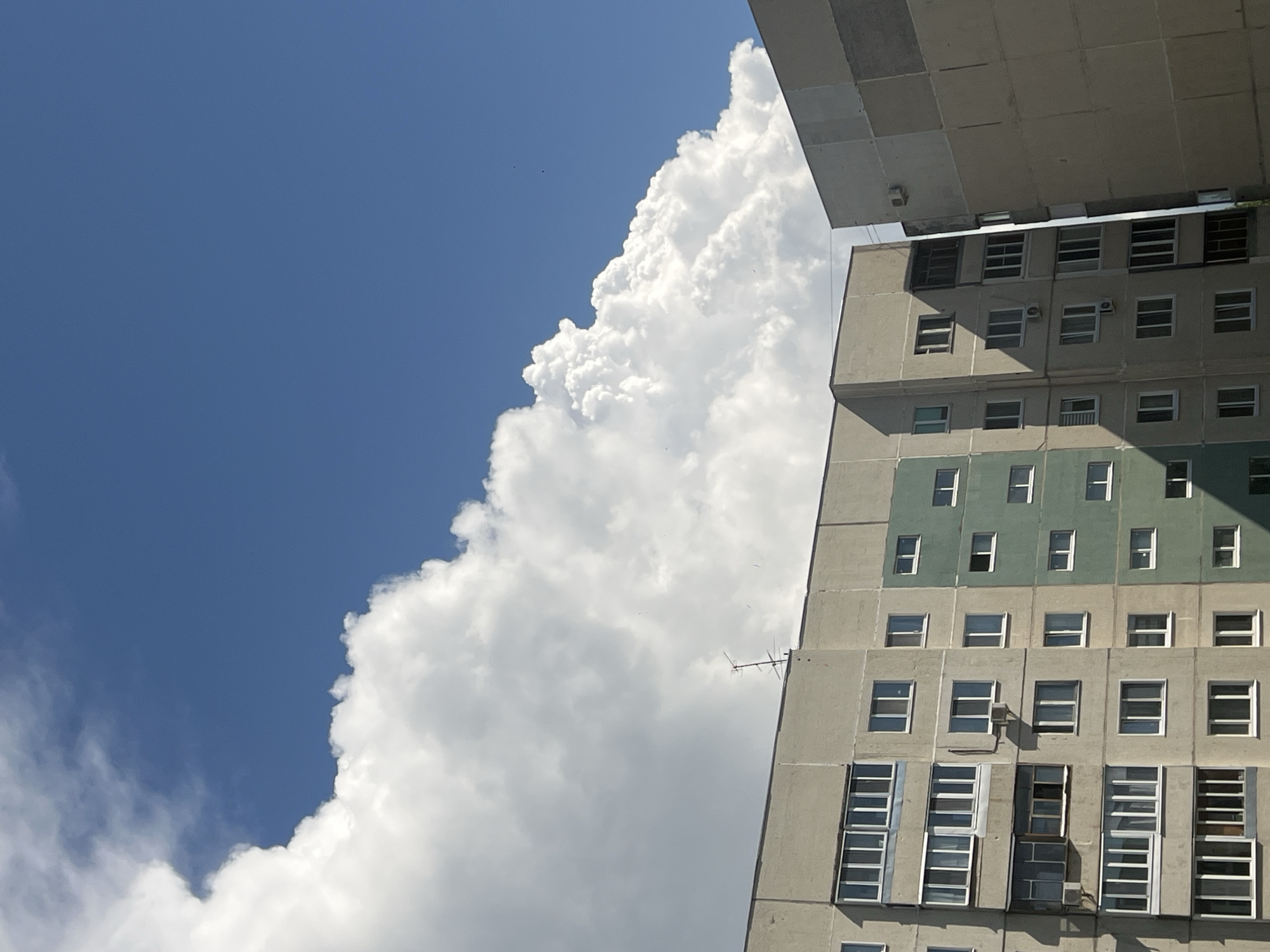 The image depicts a segment of the Ulyanovsk, Russia urban landscape, featuring the upper portions of two adjacent apartment buildings against a backdrop of a bright blue sky filled with large, white, cumulus clouds. The buildings are constructed from concrete and appear to be of Soviet-era architecture. One building is light gray with multiple windows, some featuring balconies, and a section painted green. The other building, partially visible on the right, is also concrete-colored. An antenna is visible on the roof of the left building. The lighting suggests a sunny day, with shadows cast on the buildings. There are no people or visible actions.