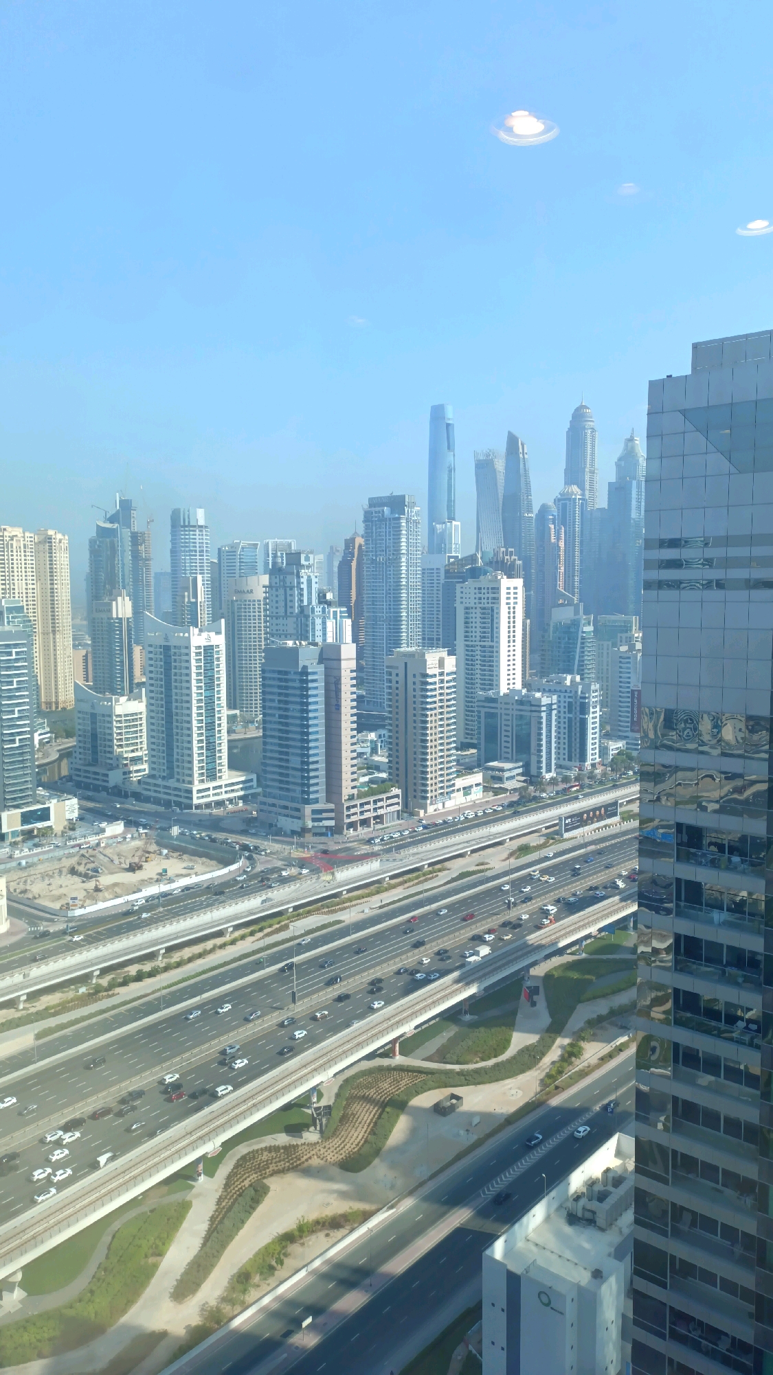 The image captures a panoramic view of a modern cityscape in Dubai, United Arab Emirates, likely from a high vantage point. The scene is dominated by numerous skyscrapers, characterized by their contemporary architectural designs and reflective glass facades. These buildings are clustered together, forming a dense urban environment.
A prominent feature of the image is a multi-lane highway that cuts through the city. Numerous vehicles, including cars and trucks, are visible on the highway, indicating active traffic flow. The highway is elevated in some sections, with overpasses and bridges supporting its structure. Adjacent to the highway, there are landscaped green spaces and construction sites, suggesting ongoing development and urban planning.
The sky is a clear blue, with some diffused light, indicating it is daytime. The overall impression is one of a bustling, advanced, and rapidly developing metropolitan area. The foreground is partially obscured by the reflective surface of a nearby building, which mirrors the surrounding cityscape and the sky.