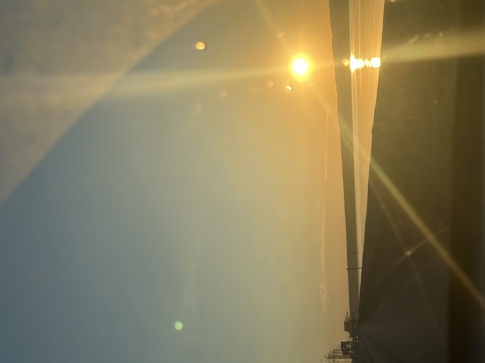 The image depicts a sunset over the Kama River in Chaikovsky, Russia. The sun, a bright orb, is low on the horizon, casting a warm, golden glow across the water and the sky. The sky transitions from a pale blue at the top to a deep orange near the horizon. A faint reflection of the sun shimmers on the water's surface. In the foreground, a dark, paved road stretches into the distance, with the silhouette of a body of water to the right. Along the left side of the road, there are several utility poles and structures, suggesting a developed area. There are no people visible in the image. The overall scene is serene and tranquil, capturing the natural beauty of a sunset over a large body of water.
