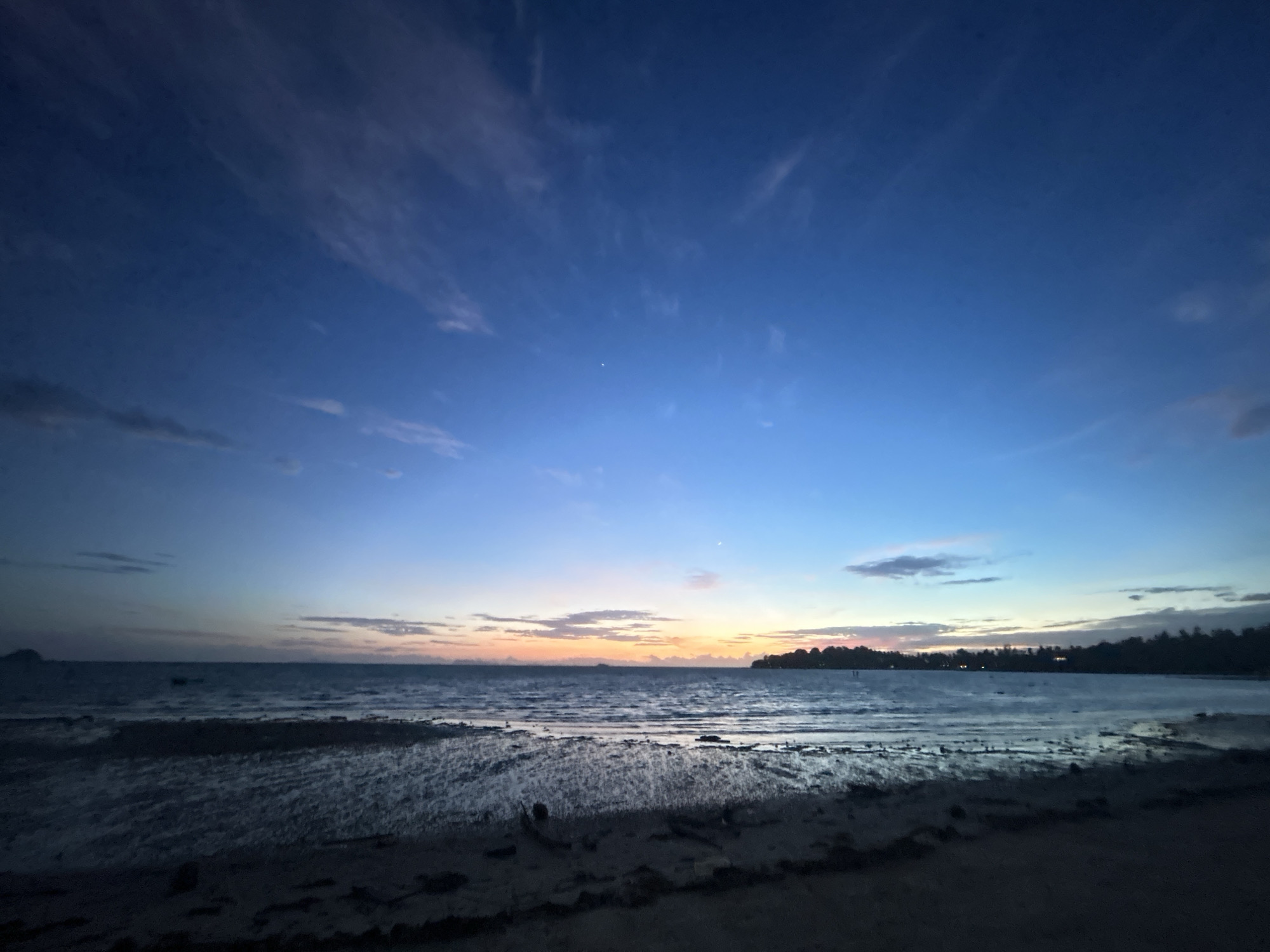 The image depicts a tranquil twilight scene at a beach on Ko Pha-ngan District, Thailand. The sky transitions from a deep blue overhead to soft hues of orange and pink near the horizon, where a faint sunset is visible. Wispy clouds are scattered across the sky. The sea is calm, with gentle waves lapping at the shore. The foreground consists of a sandy beach, with some wet sand reflecting the dim light. In the distance, a dark, tree-lined coastline is visible, with a few lights suggesting human habitation. There are no people or specific objects clearly identifiable in the scene. The overall impression is one of peacefulness and natural beauty at dusk.
