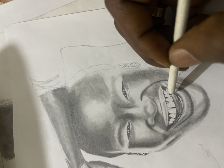 The image displays a close-up, overhead view of a graphite pencil drawing on white paper. The drawing is a detailed portrait of an individual, likely of African descent, depicted with a broad smile, mouth open, revealing a full set of teeth with noticeable gaps. The facial features are rendered with varied shading, suggesting a dark skin tone, and convey an expression of joy or laughter. In the lower right quadrant, a dark-skinned hand, wearing a metallic ring on the ring finger, holds a white blending stump and is actively applying it to refine the shading around the teeth of the drawn subject. Faint pencil marks and shading tests are visible in the upper left corner of the paper. The immediate environment indicates an indoor workspace. The precise location within Jos, Nigeria, is not visually identifiable in the image.