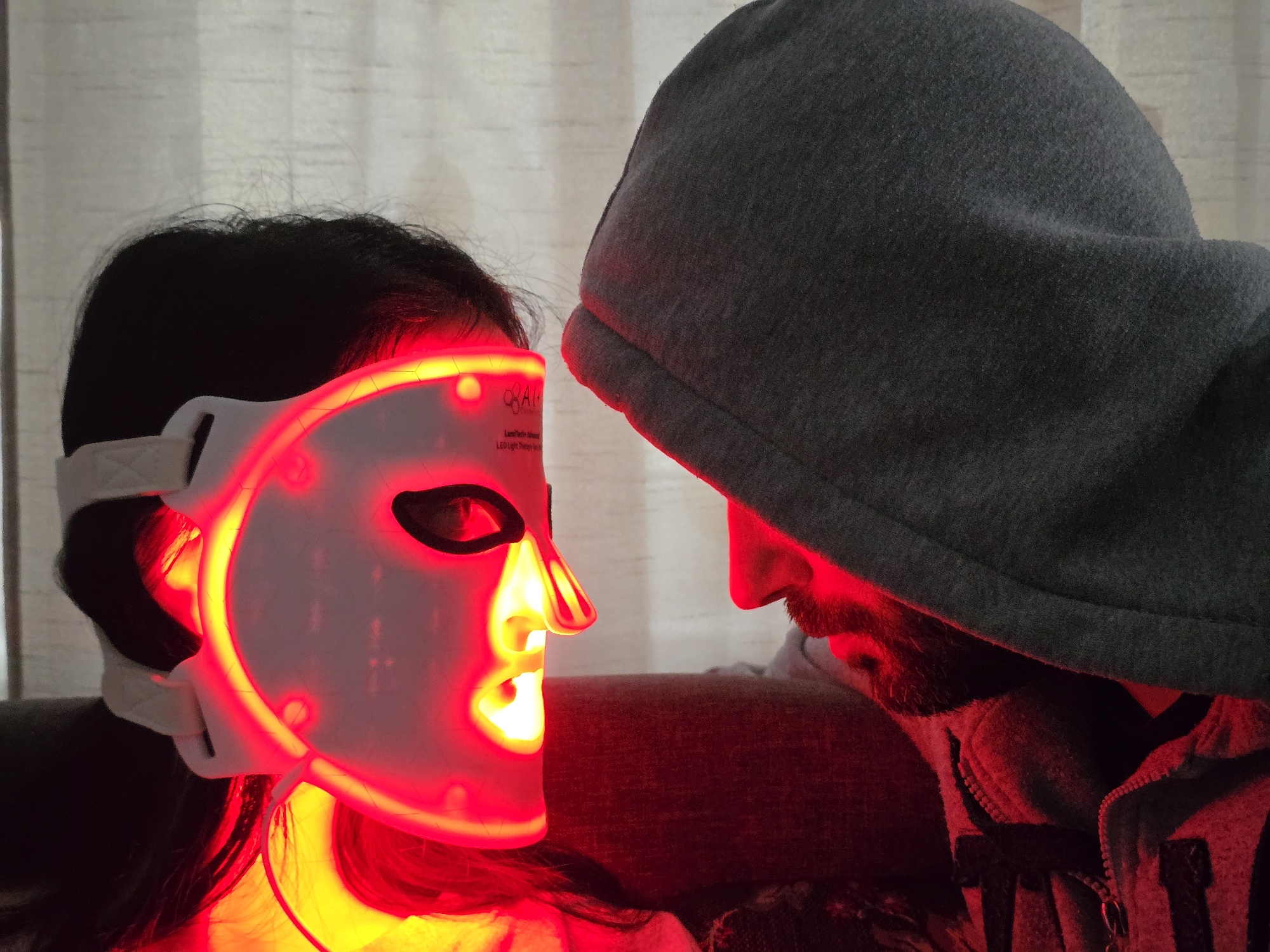 A woman wearing an illuminated red LED facial mask stands close to a hooded individual. The woman's mask is white with eye and mouth openings, and a strap securing it to her head. The hooded figure is wearing a gray hooded sweatshirt, the hood obscuring most of the face except for the lower part, including the nose, mouth, and beard. The red light from the mask casts a strong glow on both subjects. The background features a blurred curtain. The city and specific location are unknown.