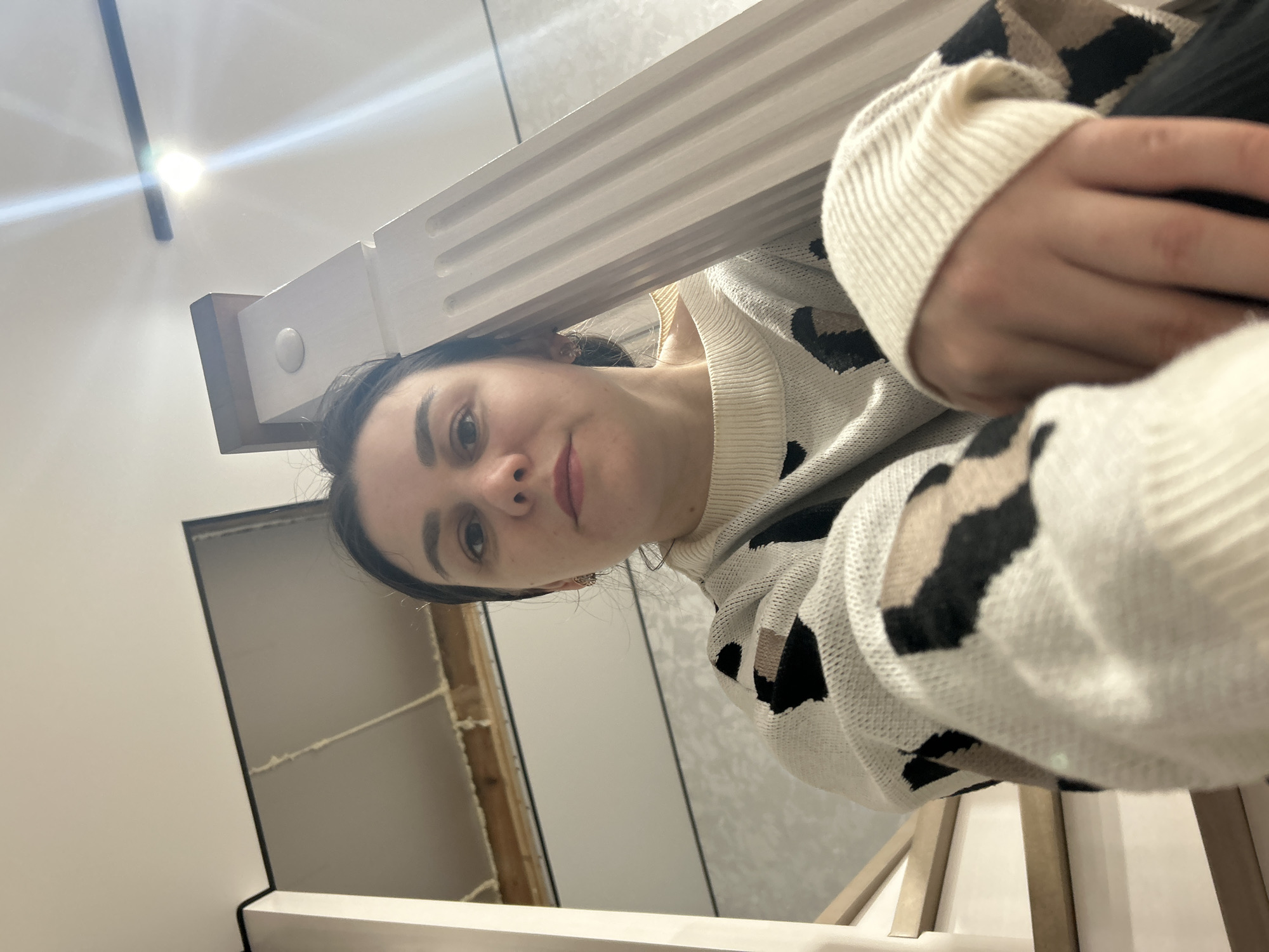 A woman with dark hair is looking down at the camera. She is wearing a white sweater with a black and tan leopard print. She is positioned under what appears to be a white bed frame. In the background, there is a wall that looks like it is under construction, with exposed wood framing. The location is unknown and cannot be determined from the image.