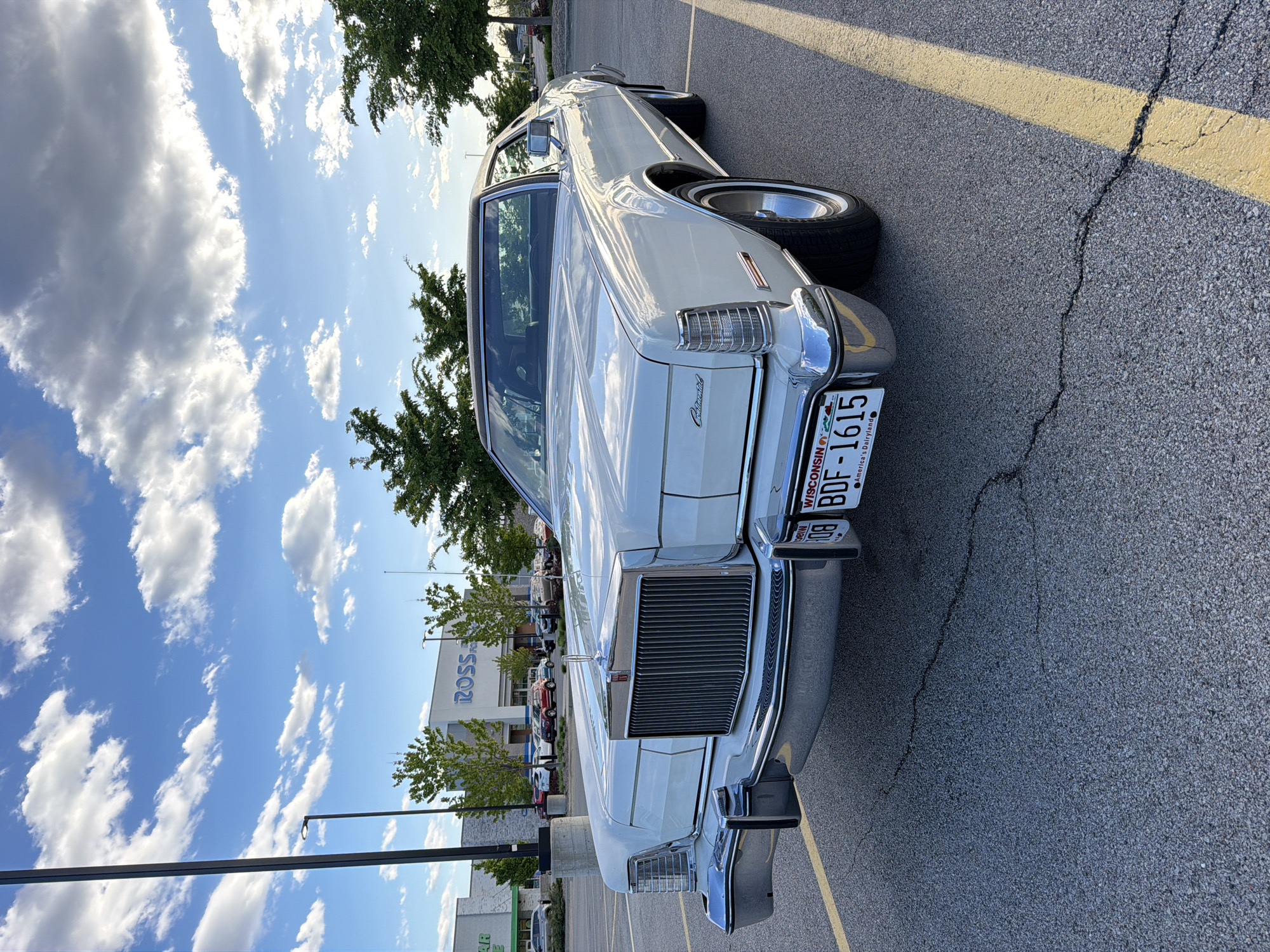 The image shows a white Lincoln Continental car parked in a parking lot in Morton Grove, Illinois, United States of America. A "Ross" department store is visible in the background. The car has a Wisconsin license plate numbered BDF-1615. The sky is blue with scattered white clouds. There are trees in the background near the store. The parking lot surface is asphalt with yellow painted lines. A light pole is visible near the parked car.