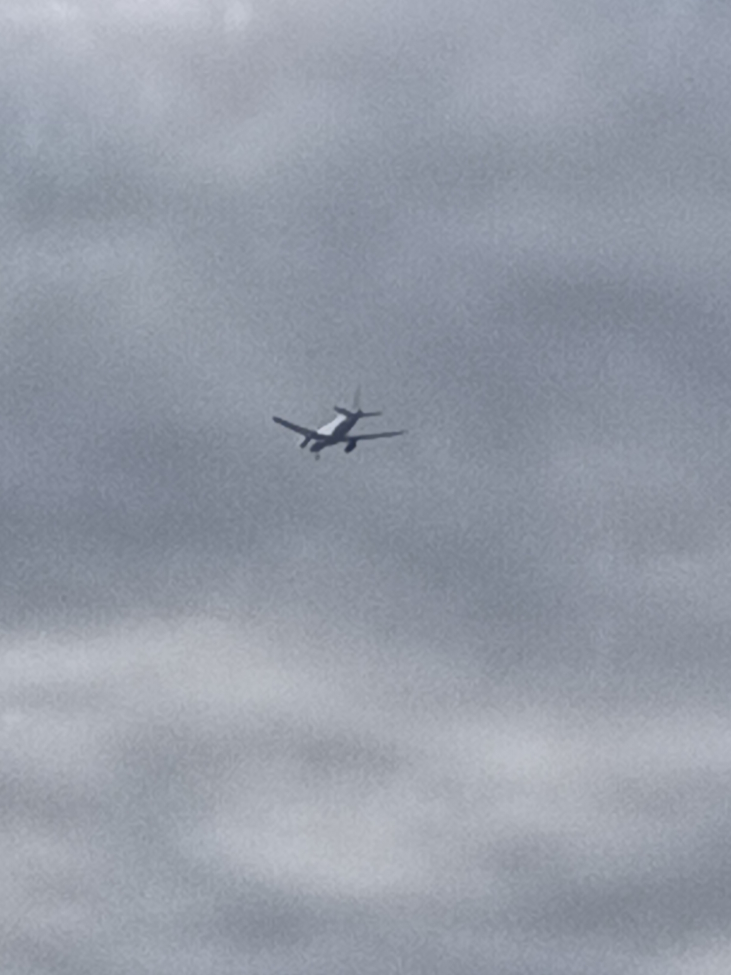 A small, propeller-driven airplane is captured in mid-flight against a hazy, overcast sky. The aircraft is silhouetted against the clouds, with its wings and tail visible. The overall impression is one of a quiet, subdued atmosphere, typical of a cloudy day. There are no discernible landmarks or urban features to indicate a specific location within Prague, Czechia, or any other city. The image focuses solely on the airplane and the sky.