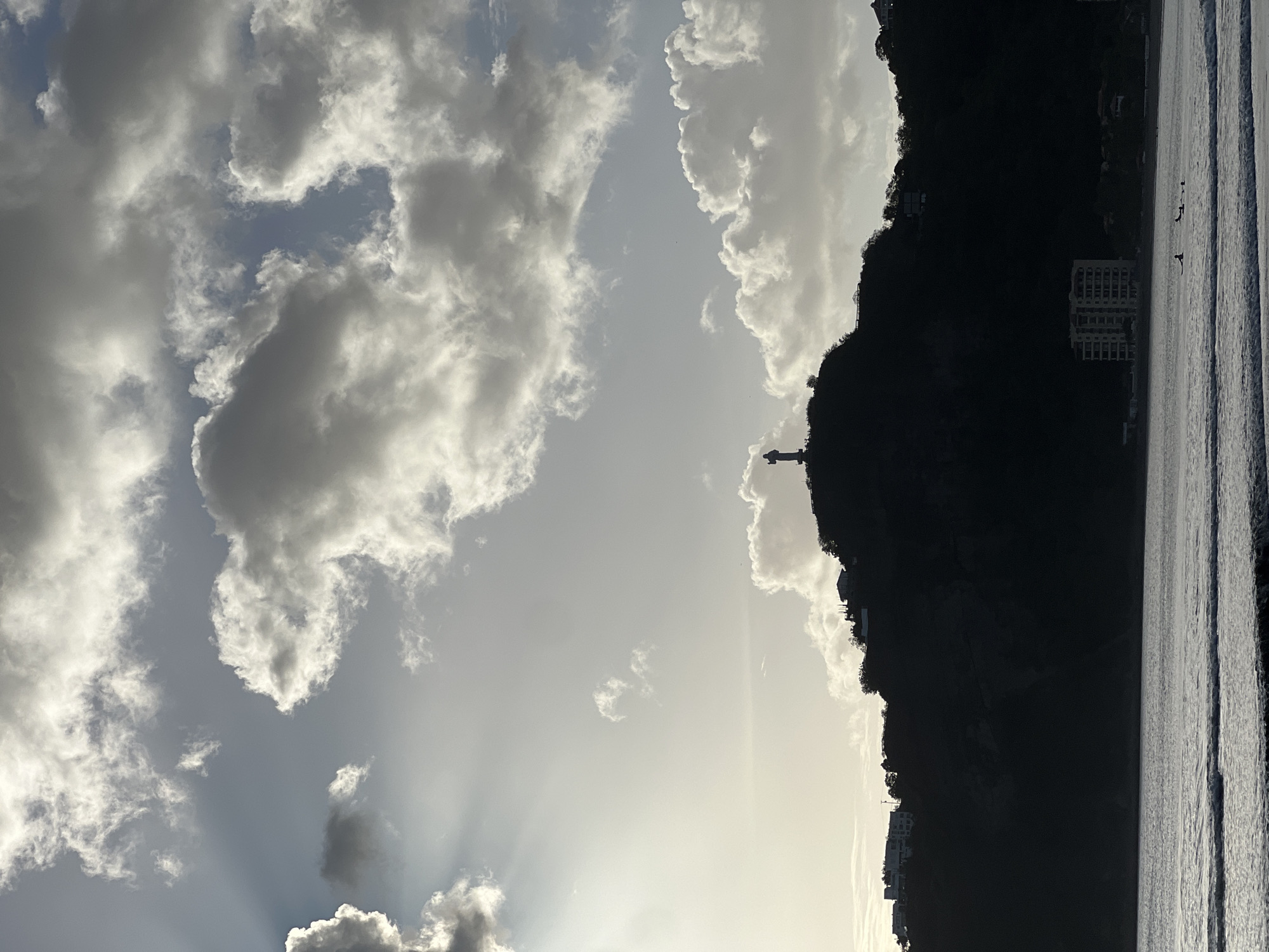 The image depicts a silhouette of the Cristo de la Misericordia statue atop a hill overlooking the bay of San Juan del Sur, Nicaragua. The statue is positioned on the right side of the hill's crest, facing towards the sea. The sky is filled with dramatic clouds, with sunbeams piercing through on the left side of the frame. Below the hill, a dark silhouette of the coastline is visible, with several buildings and structures dotting the hillside. The foreground features the calm waters of the bay with small waves gently lapping the shore. A few surfers are scattered in the water. The overall scene is a dramatic and serene coastal landscape.