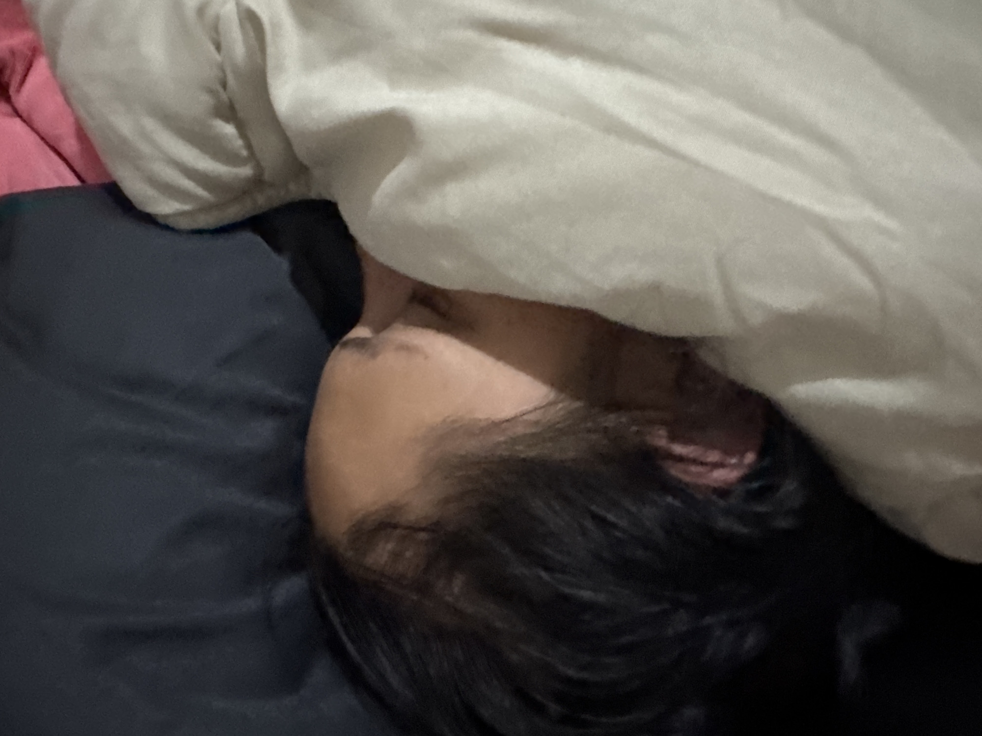 A person with black hair is sleeping in bed with a white blanket partially covering their face. The bed has a black sheet, and a pink blanket is visible near the person's head. There is no information to suggest the location is Sofia, Bulgaria.