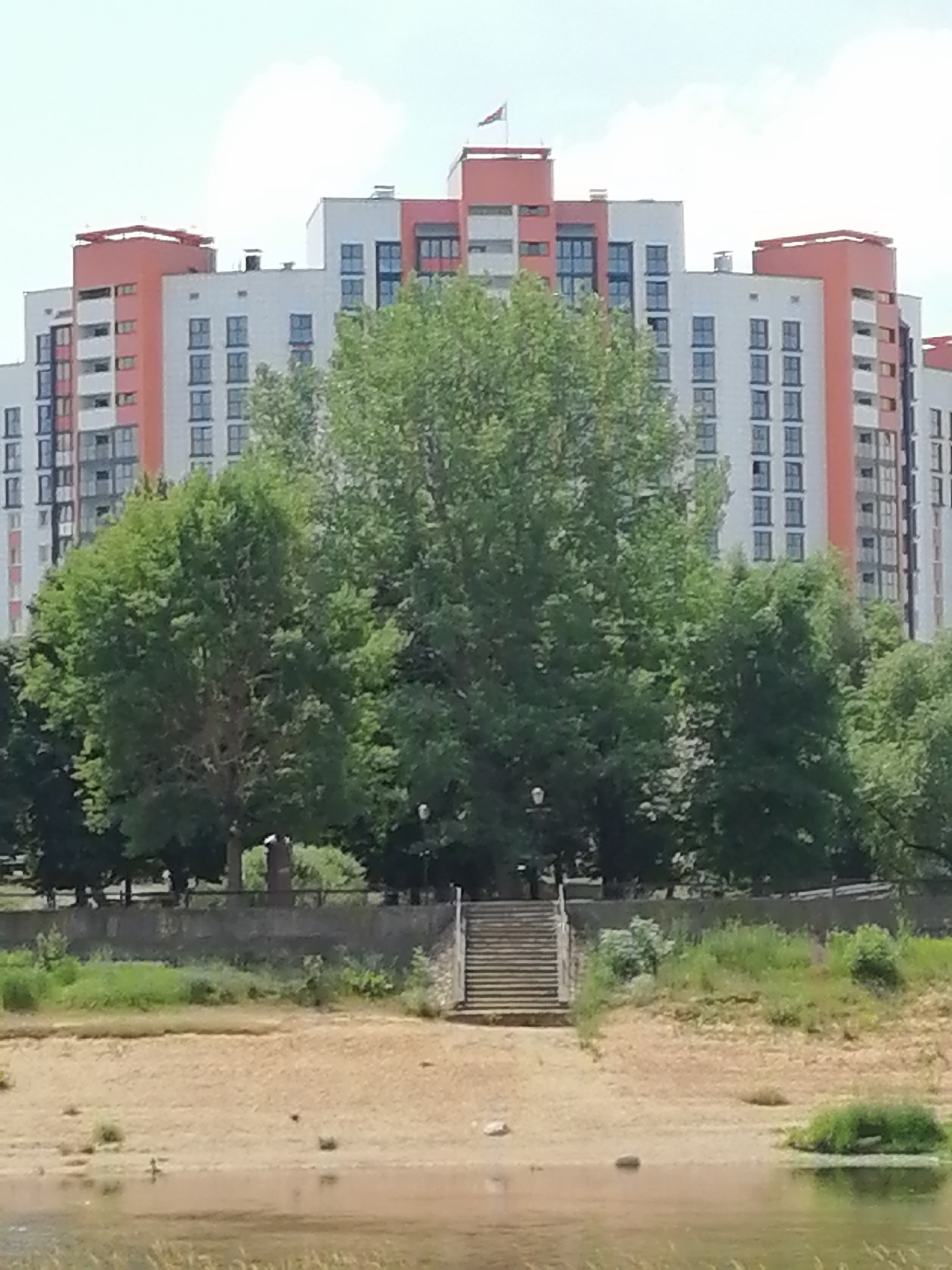 The media file shows a wide shot of a modern, multi-story residential building complex situated behind a line of lush green trees. The buildings are tall and feature a contemporary design with alternating sections of white and orange facades. Numerous windows are visible on each floor, suggesting many apartments. A flag is flying from the top of one of the buildings.
In front of the buildings, a dense canopy of large trees dominates the mid-ground, obscuring much of the lower portions of the structures. The trees appear full and healthy, indicating it is likely summertime.
The foreground consists of a sandy embankment leading down to a body of water, possibly a river or lake. The embankment is partly covered with dry grass and scattered rocks. A set of concrete stairs is visible on the right side of the embankment, providing access to the area above. A wall or railing runs along the top of the embankment, separating it from the trees and buildings.
The sky is light and somewhat hazy, with scattered clouds. The overall lighting suggests it is daytime, likely late morning or afternoon, under fair weather conditions. There are no people visible in the scene, and the environment appears to be a public space or park area adjacent to the residential complex. The scene evokes a sense of urban development integrated with natural elements. No legible text is present in the image.