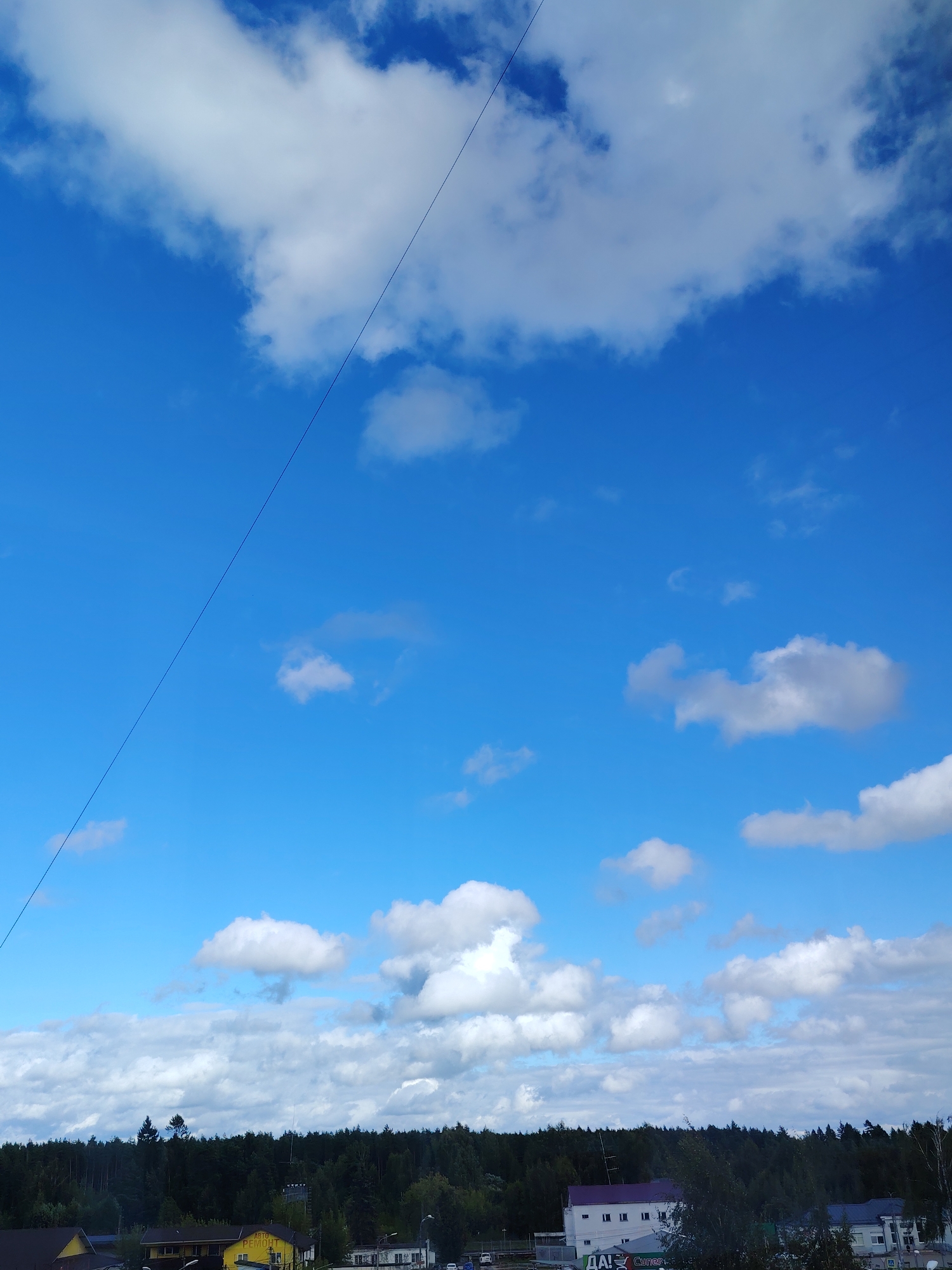 This image captures a daytime scene in the city of Konakovo, Russia, under a bright blue sky dotted with white, fluffy clouds. A thin, dark line, likely a wire or cable, diagonally traverses the sky from the upper left to the lower right. In the background, a dense line of dark green trees forms a natural border. Below the trees, several buildings are visible. One prominent yellow building has Cyrillic text that appears to read "АВТО РЕМОНТ" (Auto Repair). Adjacent to it are other structures, including a white building with a purple roof. The overall impression is of a semi-rural or suburban area with natural elements and functional buildings.
