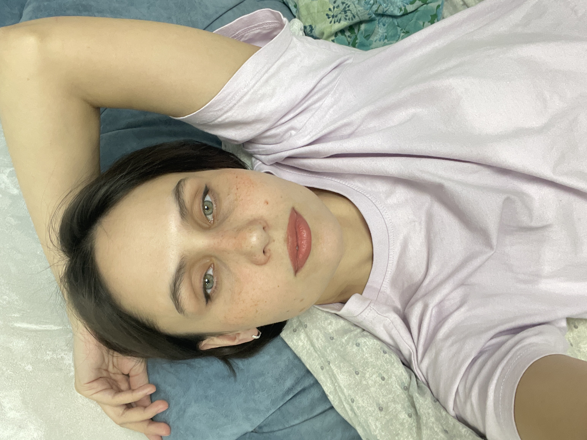 A young woman with fair skin and freckles lies on her back, looking at the camera. She is wearing a light purple t-shirt. Her hair is dark and short. She has light green eyes and her lips are a coral color. Her right arm is raised above her head. She is lying on a bed with a blue pillow and patterned sheets. The location is not discernable from the image.