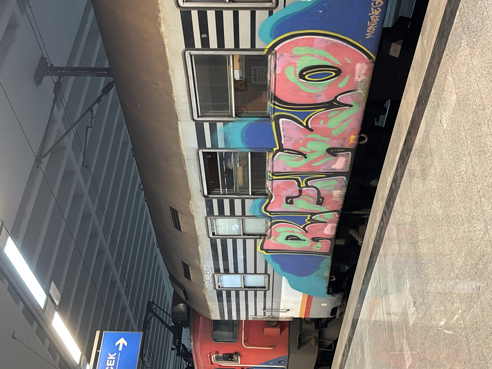The image shows a train at a station platform. The train car is painted with graffiti and has the word "DENKO" prominently displayed. Above that is written the word "MONTE NEGPCG" which suggests the train may operate in Montenegro. The train car is white with black stripes. A red locomotive is visible at the front of the train. A sign with a blue background and white arrow points in the direction of "CEK". The platform is made of stone and there is an overhanging ceiling structure. The location is likely Belgrade, Serbia, because "CEK" likely references the acronym of a bus station.
