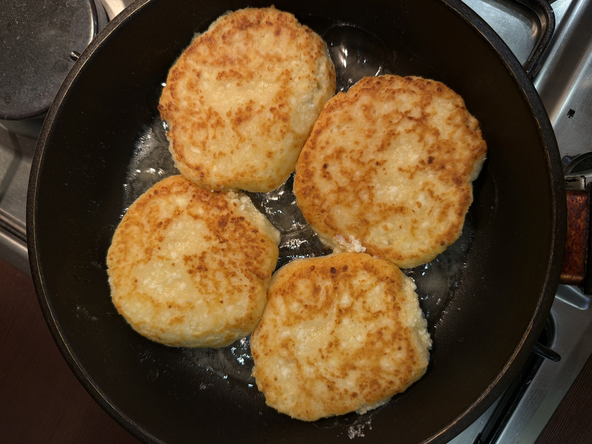 A top-down shot shows four golden-brown, circular patties cooking in a black cast iron skillet. The skillet rests on a gas stovetop. The stove's surface is silver, with visible metal parts. A dark wooden surface is also visible in the lower left corner.