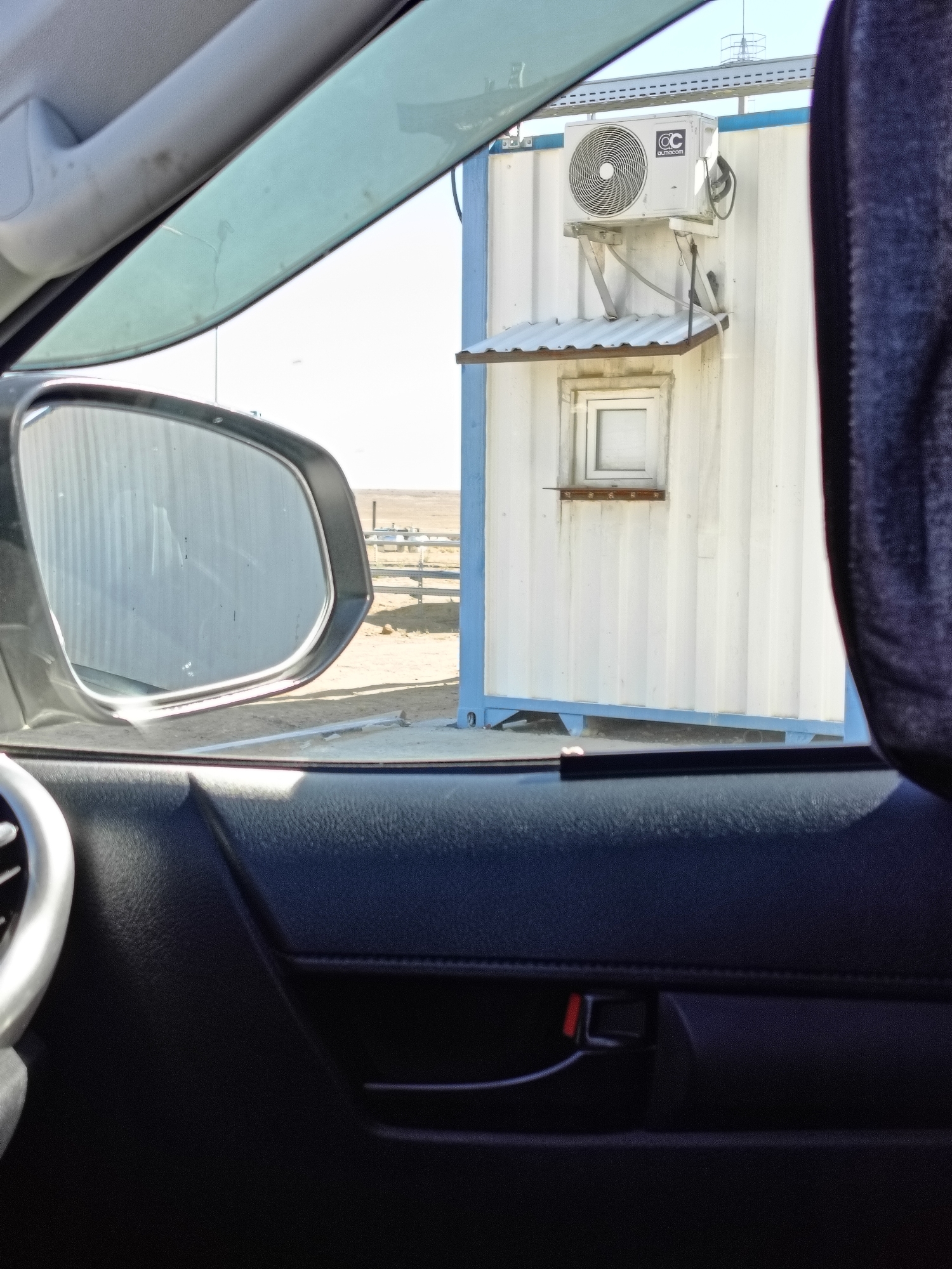 The image captures a view from inside a vehicle, looking out through the side window and side mirror. In the foreground, a portion of the car's interior is visible, including the door panel with a black leather-like texture and a silver vent on the left side. The side mirror reflects a corrugated metal wall.
Beyond the vehicle, the scene opens up to an outdoor environment. A white, rectangular portable building, possibly a shipping container converted into a small structure, stands prominently. It has a blue base and a light blue trim along its roofline. An outdoor air conditioning unit is mounted on the upper part of the building. Below the air conditioner, there is a small, white-framed window with a simple metal awning over it. The ground outside appears to be dusty and dry, with some low-lying vegetation and fencing in the background, suggesting an arid or semi-arid landscape. The sky is bright and clear, indicating daytime. The image has a slightly washed-out quality, possibly due to bright sunlight. The location is identified as Unknown, Kazakhstan.