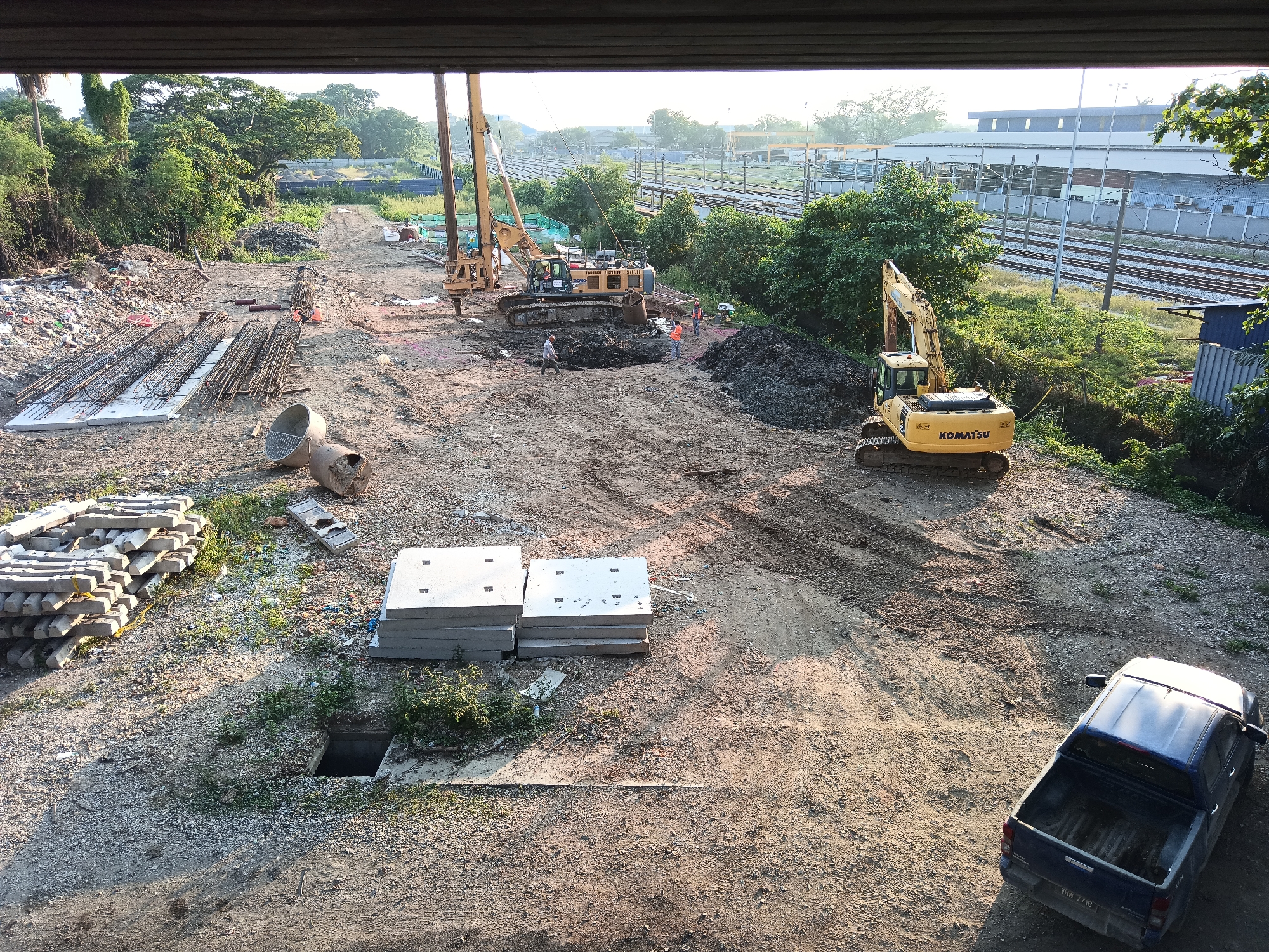 The image depicts a construction site in Klang City, Malaysia, near a railway line. A large yellow drilling rig with a long mast is positioned in the center of the frame, with its boom extended. In front of the drilling rig, a yellow excavator is parked, and a pile of dark soil is situated to its right. Several construction workers, dressed in orange vests and helmets, are visible near the drilling rig and the excavator.
To the left of the drilling rig, a pile of construction materials, including rebar cages and concrete slabs, is stacked on the ground. Several cylindrical concrete structures are also present. Further left, a large pile of debris, including plastic waste and construction remnants, is visible.
In the background, a railway line runs parallel to the construction site, with several tracks and overhead electrical infrastructure. A modern building with a light-colored facade is situated beyond the railway.
To the right of the scene, another yellow excavator is actively working, digging into a mound of earth. In the foreground, a dark blue pickup truck is parked. The ground is predominantly dirt and gravel, with tire tracks indicating recent vehicle movement. The overall environment suggests an active construction zone undergoing development or maintenance, likely related to infrastructure work near the railway.