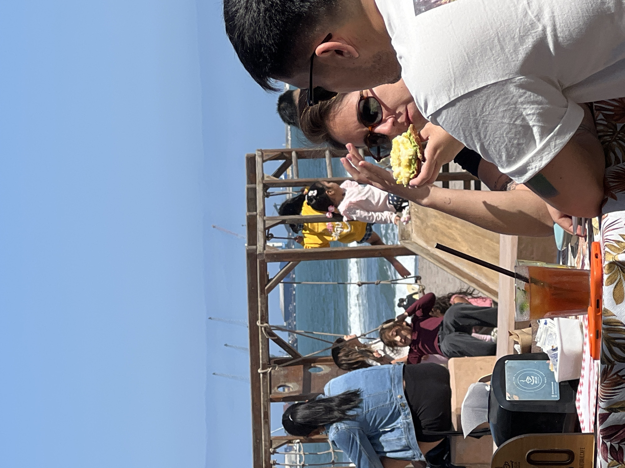 The image shows a group of people at a restaurant in Arica, Chile, near the coast. The foreground captures a couple eating and drinking at a table covered with a tropical-print tablecloth. A drink with a straw is visible. In the background is a wooden play structure, with several children and adults interacting with it. The ocean and clear blue sky are visible behind the play structure. The focus seems to be on the foreground subjects, but the environment suggests a casual, outdoor dining experience with recreational activities nearby, possibly at a waterfront park or restaurant with a playground.
