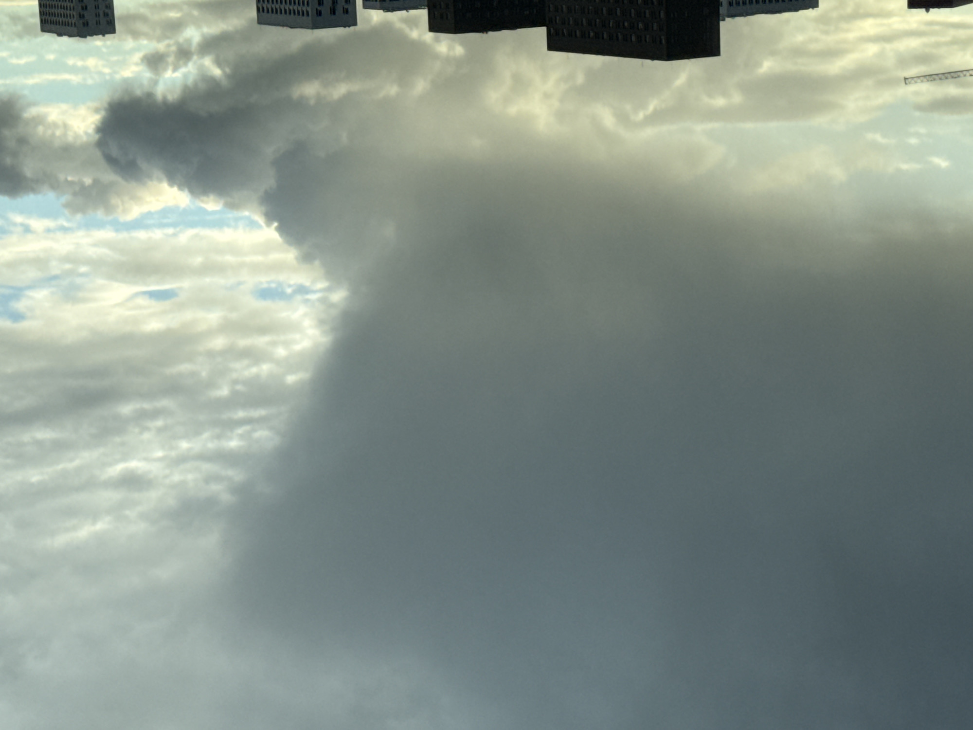 The image, oriented upside down, depicts a cloudy sky above what appears to be a cityscape. Several buildings are visible along the top edge of the frame. A large mass of dark, gray cloud occupies a significant portion of the image, contrasting with lighter clouds and patches of blue sky. A crane is also visible. No people or specific interactions are discernible. The location is presumed to be Khimki, Russia, though this cannot be confirmed by visual evidence alone.