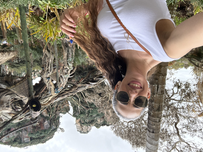 An adult female is depicted taking a selfie outdoors in Pandora – The World of Avatar at Disney's Animal Kingdom, located in Bay Lake, Florida, United States. The subject, in the foreground, has long, wavy, reddish-brown hair, wears hexagonal-framed sunglasses, a white sleeveless top, and a gold-colored necklace with an infinity symbol pendant. She is smiling at the camera, with her right hand, displaying pink and white polka-dotted fingernails, touching her hair. The background consists of intricate, themed environmental structures characteristic of Pandora, featuring large, gnarled trees, exposed root systems, and simulated "floating mountains" covered in lush green foliage. Several dark, flute-like architectural elements with straw caps and glowing yellow interiors are integrated into the scene. A tall, bare tree trunk stands on the left. The ground is covered with various green plants. A partially visible figure in blue is in the distant background. The sky is overcast.
