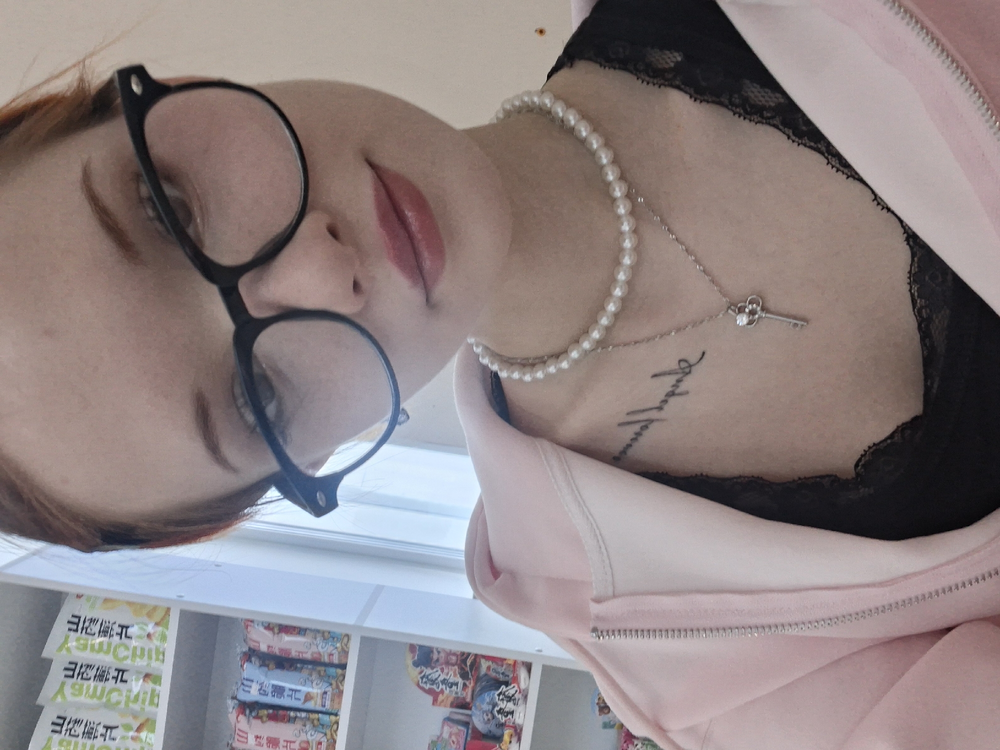 A woman with glasses and a pink sweater is indoors in Chaikovsky, Russia. She is wearing a pearl necklace with a key pendant and a black lace top. She has a tattoo on her chest. Behind her is a shelf containing various packaged goods.