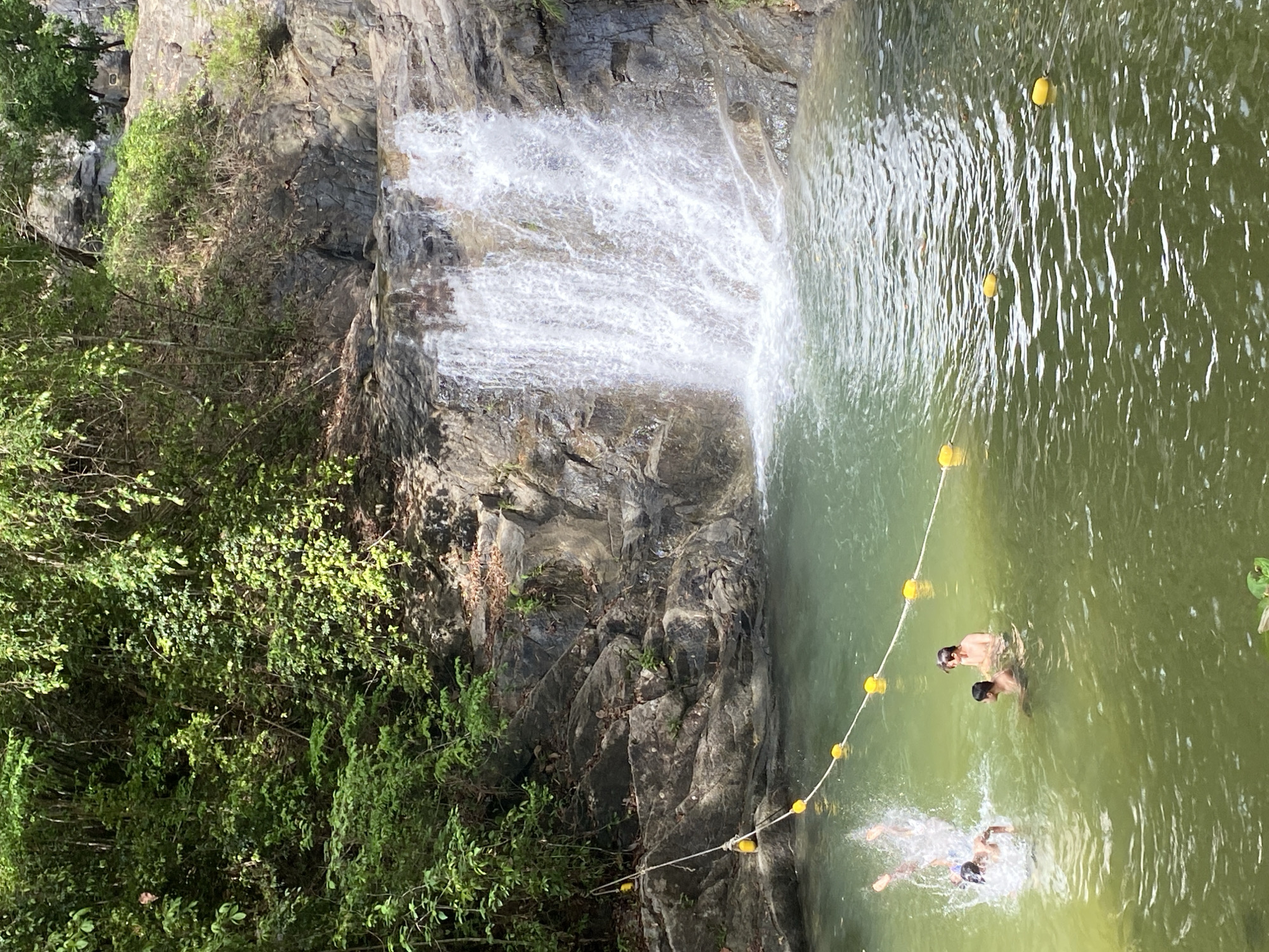 The image shows a waterfall cascading into a pool of green water, likely located in a natural area. The waterfall descends over a rocky cliff face partially covered with vegetation. A safety line with yellow buoys stretches across the pool. Three individuals are in the water: one swimmer is submerged and splashing, while two other individuals are partially submerged, standing or treading water near the buoy line. The surrounding environment consists of lush green foliage and rocky terrain, indicative of a natural, possibly mountainous location. The scene is captured in daylight. Based on the prompt, the location is likely in Tambon Thai Mueang, Thailand.
