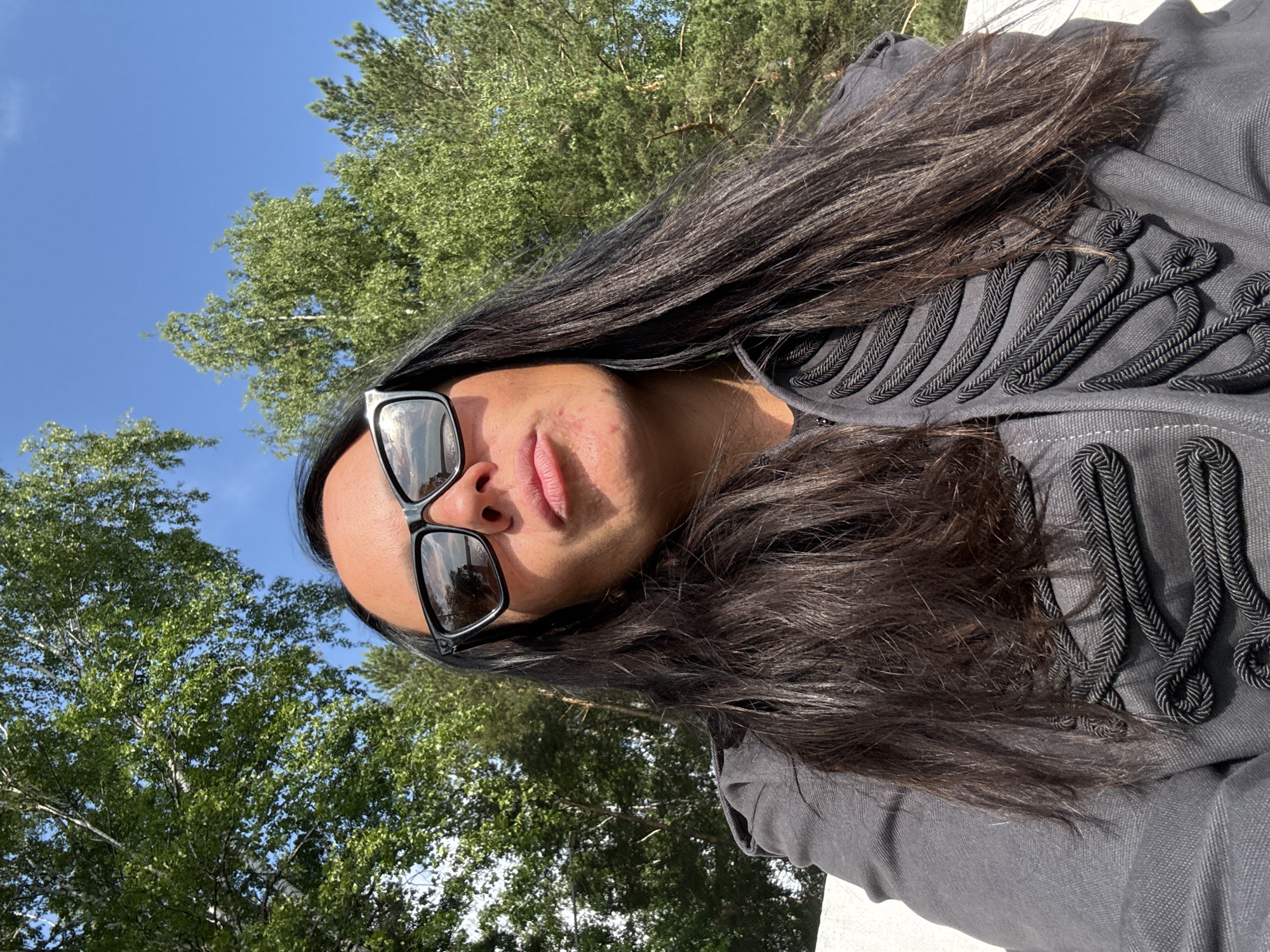 A low-angle, medium shot captures a woman wearing sunglasses and a gray jacket with elaborate black braided trim across the chest and shoulders. She has long, dark hair that falls around her face and shoulders. The woman is outdoors with a backdrop of green trees and a clear blue sky. The scene appears to be in Chaikovsky, Russia.