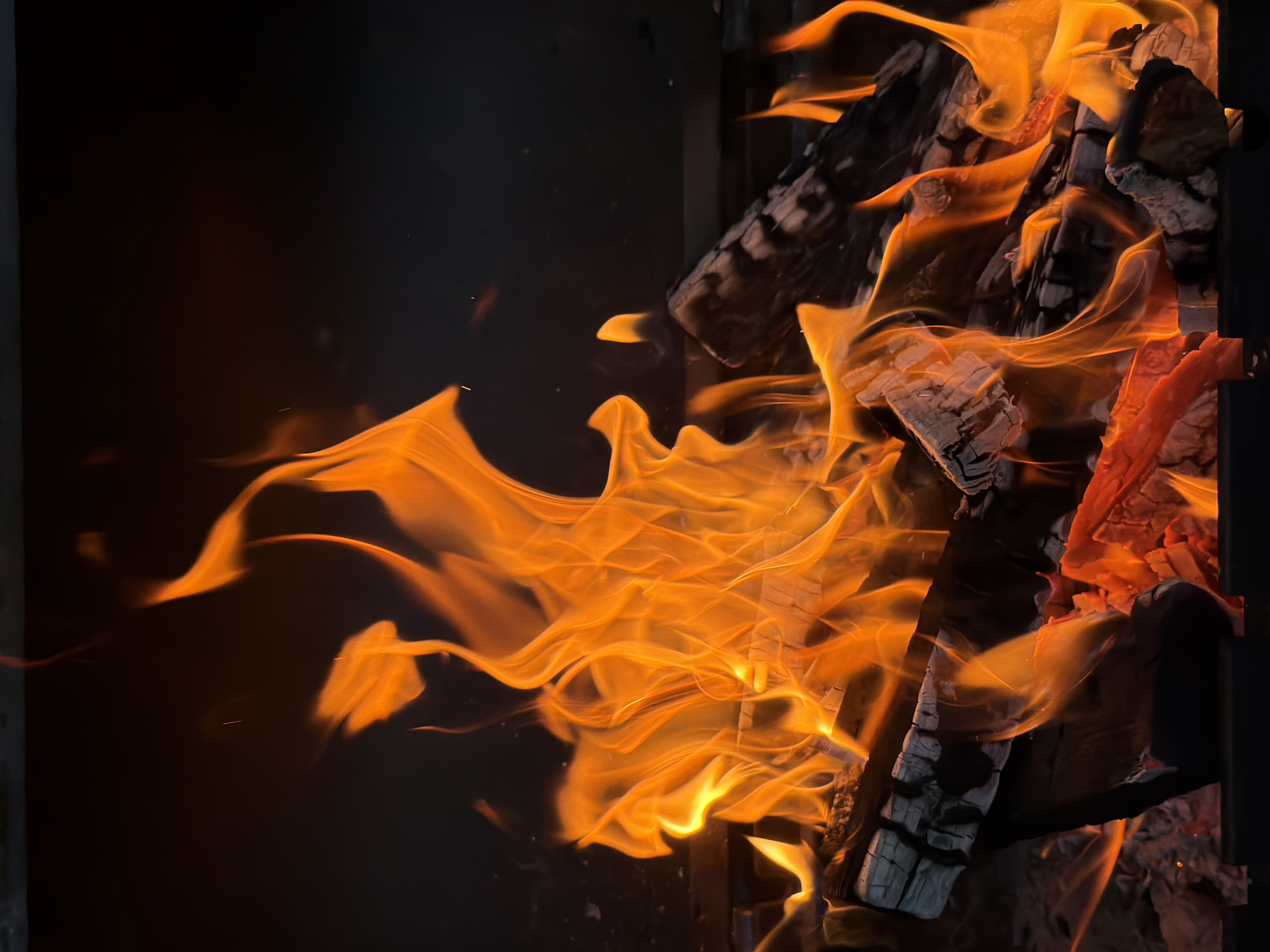 A close-up, eye-level shot captures a vibrant campfire. The flames, rendered in intense oranges and yellows, leap and dance above glowing embers and charred logs. The logs, some dark and ashy, others revealing hints of red-hot interior, are stacked haphazardly within a dark, metallic structure, likely a fire pit. The background is a deep, indistinct black, providing a stark contrast that makes the fire's brilliance all the more dramatic. The scene is devoid of people or any other distinguishing features, focusing solely on the dynamic and primal energy of the burning wood.