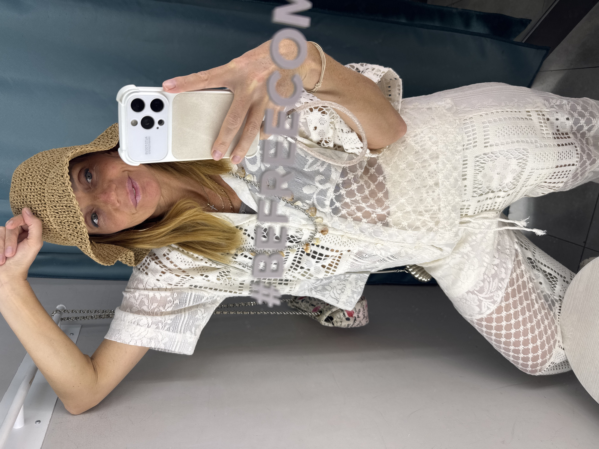 A woman is taking a selfie with a smartphone in what appears to be a fitting room. She is wearing a straw hat and a white lace outfit. The fitting room has a blue curtain. The location in the city of Saint Petersburg, Russia, is not identifiable from the image.