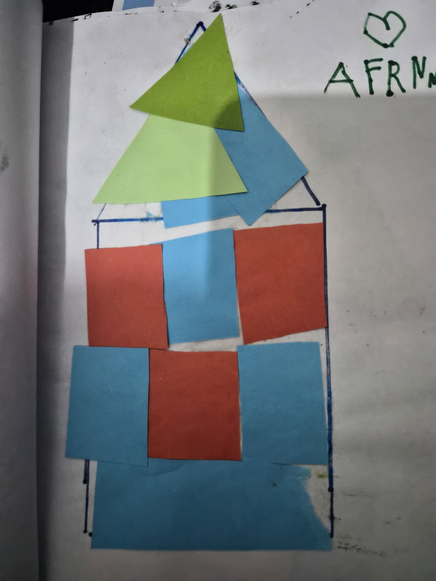 The image is a close-up, overhead shot of a piece of paper with a collage of colored paper shapes forming a house. The paper is white and appears to be part of a notebook or sketchbook, with a curved edge on the left side suggesting it's from a spiral-bound book. The house is constructed with red and blue squares for the body and light green and blue triangles for the roof. The outline of the house is drawn in black marker. In the upper right corner, there is a green heart shape and the letters "AFRN" written in black marker. The background is a white paper surface with some smudges and faint lines. There are no people or animals visible in the image, and no discernible elements that would indicate a location in Balikpapan, Indonesia. The image appears to be a child's art project.