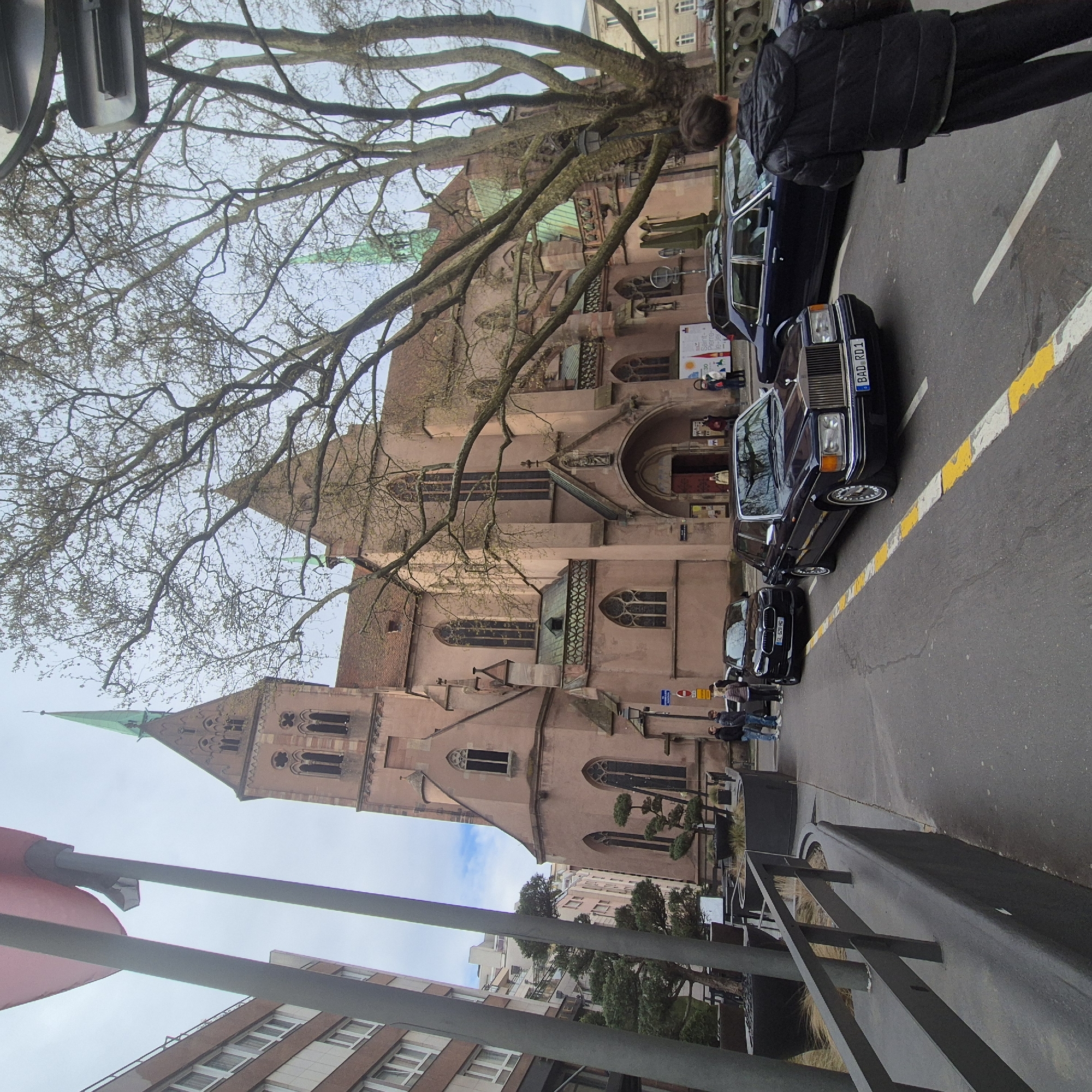 The image depicts the Saint Pierre le Vieux Church in Strasbourg, France, viewed from street level. The church's steeple, constructed from red sandstone, rises into the sky. A bare-branched tree dominates the upper part of the frame, partially obscuring the church's facade. Several black luxury cars are parked along the street, with a person in a black jacket standing next to the vehicles. The cars have visible license plates, "BAD RD1" and "TL 626 WO". A group of people stand near the cars and the church entrance. A metal fence runs along the lower right of the image, separating the street from a lower area with landscaping and trimmed trees. Other buildings and urban structures are visible in the background.