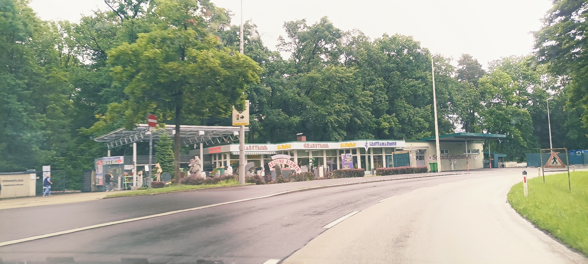 The image depicts a commercial establishment, likely a funeral service provider, situated in Linz, Austria, along a wet asphalt road. The building features a covered outdoor area supported by metal beams, displaying signage for "BLUMEN" (flowers), "GRABSTEINE" (gravestones), and "LANTIAMARMOR" (marble for graves). Various gravestones and sculptures are displayed outside the establishment. The surroundings are densely covered with lush green trees, indicating a natural or park-like setting. A pedestrian crossing sign is visible on the right side of the road, suggesting proximity to a residential or public area. Two individuals are observed near the entrance of the establishment, with one walking towards it and another standing nearby. The overall atmosphere appears overcast and slightly humid, consistent with a rainy day.
