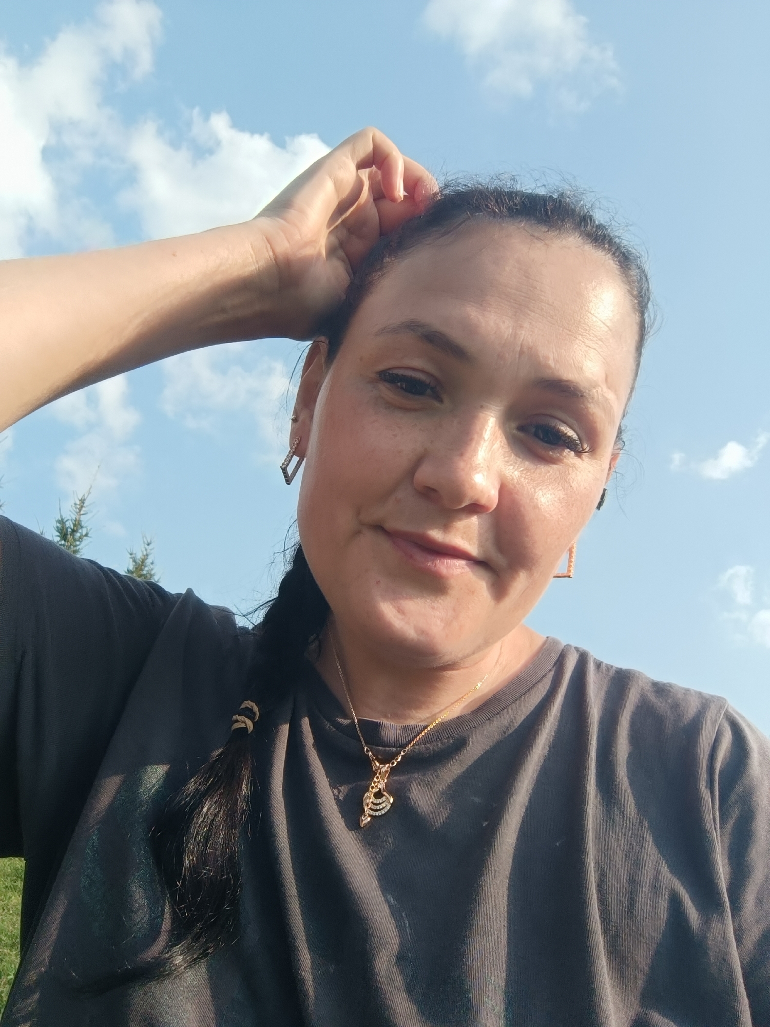 A woman is taking a selfie outdoors under a blue sky with clouds. She is wearing a dark gray t-shirt and a gold necklace with a pendant. Her hair is dark and in a braid, with a few strands loose around her face. She is touching her forehead with her right hand. The background shows trees and a grassy area. The image appears to have been taken during the daytime. The location in the city of Karsha, Russia is not identifiable from the visual information provided.