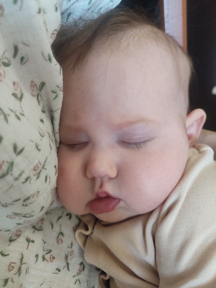 The image presents a close-up portrait of a baby peacefully sleeping. The baby is positioned on its left side, with its left cheek nestled against a soft, light-colored fabric that features a delicate pattern of small, muted green leaves and faint pink or reddish floral motifs. This fabric appears to be a blanket, swaddle, or part of a wrap, partly covering the baby's forehead and providing support.
The baby has fair skin and soft, fine hair that appears light brown or auburn, with some sparse strands on the crown of the head. Its eyes are gently closed, and subtle eyelashes are visible. The cheeks are full and rounded, contributing to a soft, cherubic appearance. The baby's small nose is upturned, and its lips are slightly parted, revealing a glimpse of the tongue, suggesting deep slumber. The lips are a natural, reddish-pink color. A small ear is partially visible on the right side of the baby's head.
The baby is wearing a simple, light-colored, long-sleeved garment, likely beige or cream, which exhibits a fine ribbed texture. The overall atmosphere conveyed by the image is one of calmness, innocence, and tranquility. The background to the right of the baby is dark and out of focus, suggesting an indoor setting. The lighting is soft and gentle, illuminating the baby's face without harsh shadows. No text is visible in the image.