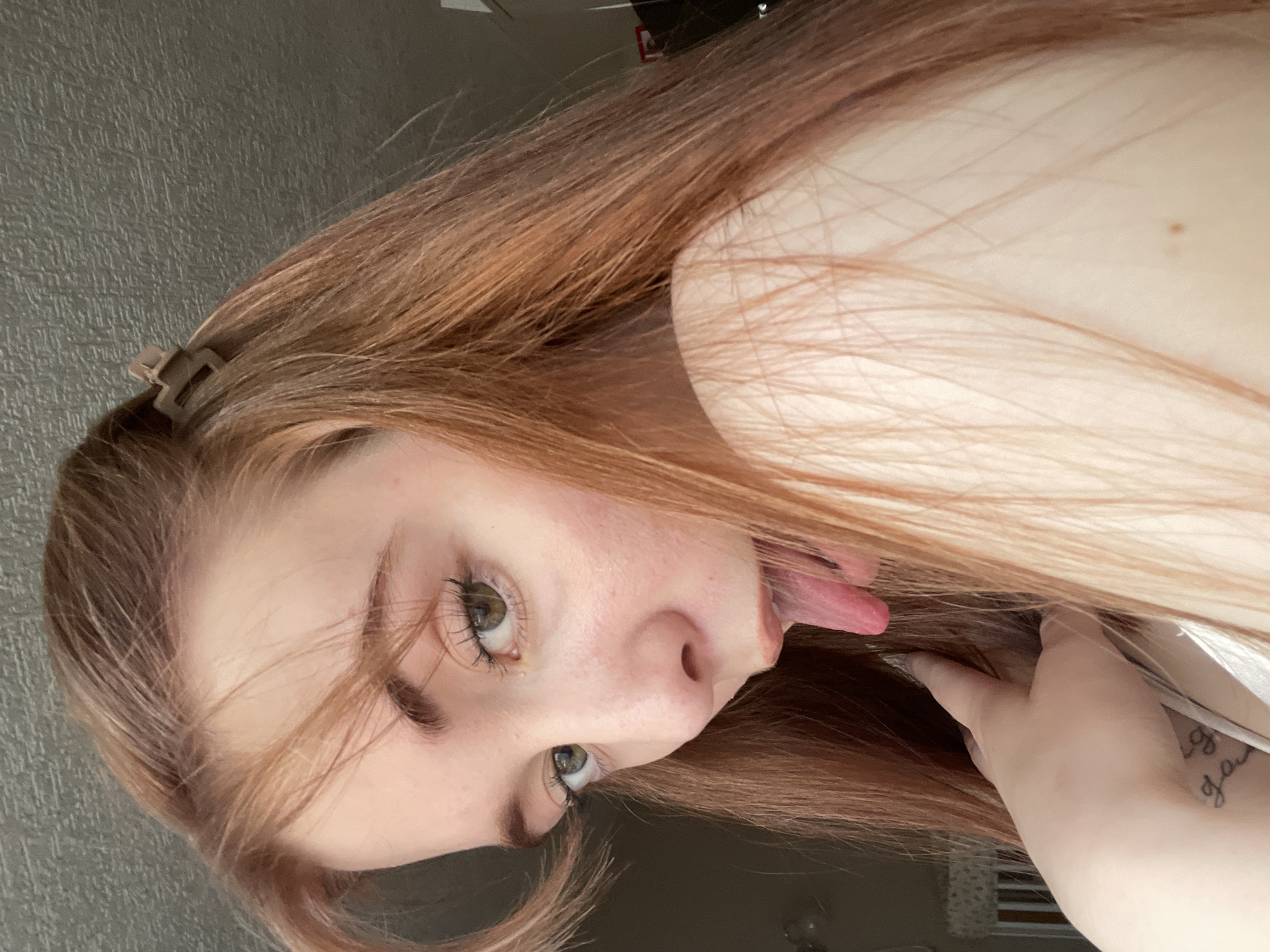 The image displays a close-up, rotated portrait of a young individual, likely female, with long, reddish-brown hair and striking green or hazel eyes. The subject is looking directly at the camera with a playful and mischievous expression, their tongue extended slightly.
Her long, flowing hair shows various shades of reddish-brown, with some strands framing her face. A small, light brown claw clip is visible in her hair on the upper left side of the frame, holding some hair back. Her eyes are accentuated with mascara.
On the right side of the image, a light-skinned hand is visible, appearing to hold or touch the subject's hair near her shoulder or neck area. On the wrist of this hand, a partial tattoo written in cursive script is discernible. The visible letters are ambiguous but appear to spell out "gory" or a similar word that is not fully legible.
The background is simple, featuring a dark grey, subtly textured wall on the left side, indicating an indoor setting. The lighting suggests daytime, with natural light illuminating the subject's face and hair from what appears to be the right side of the frame. The overall atmosphere conveyed is one of playfulness and youthful energy.