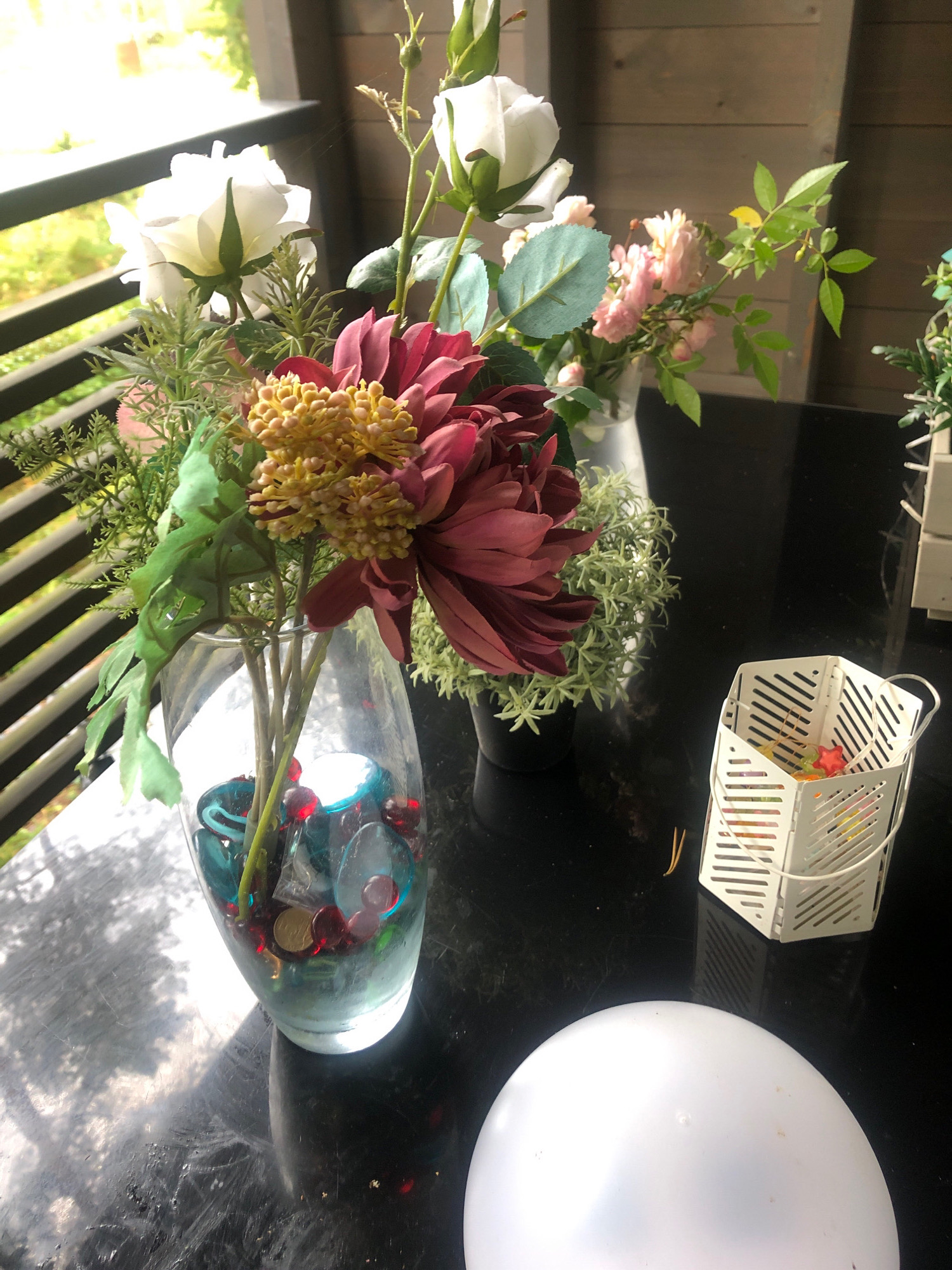 The image shows a close-up view of a black table with a floral arrangement and other decorative objects. The floral arrangement features a large, deep red dahlia with yellow accents, surrounded by smaller pink roses and white roses, all in a clear glass vase filled with blue and red decorative stones. To the right of the vase, there is a white decorative metal basket containing colorful small items, possibly candy or small ornaments. In the foreground, a portion of a white spherical object is visible. The background suggests an outdoor setting, possibly a balcony or patio, with a wooden-paneled wall and railings, and greenery visible through the railings. The overall lighting suggests it is daytime. The image does not provide any information about the city of Arfa, Russia.