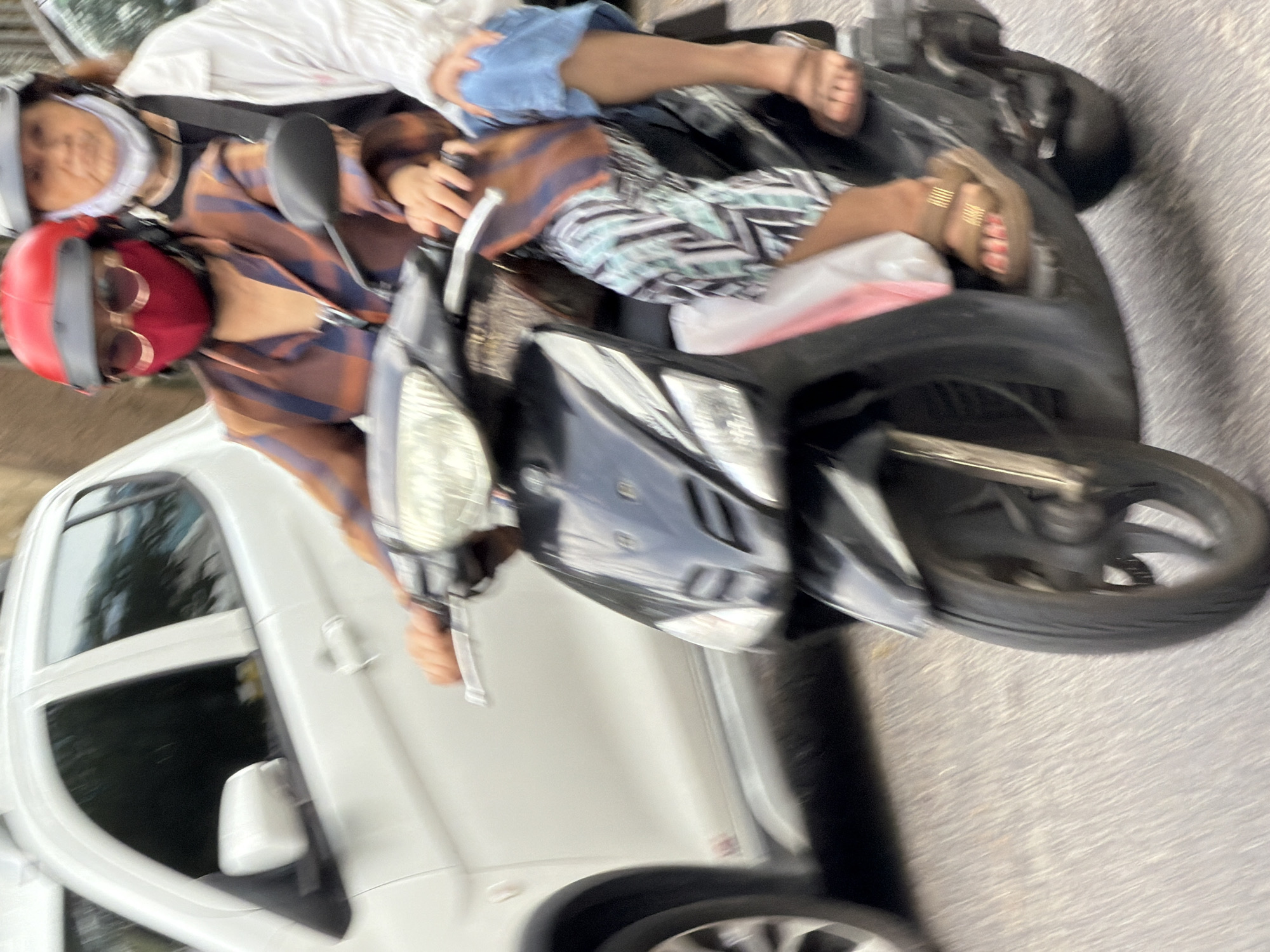 Two individuals are riding a black scooter on a paved road in Nha Trang, Vietnam. The person in front is wearing a red helmet, red mask, sunglasses, and a patterned brown and blue jacket. They are operating the scooter. The person behind them is wearing a grey helmet and a white jacket over a dark top, with blue pants. Both individuals appear to be wearing masks for protection. A white car is visible to the left of the scooter. The road surface is grey and appears to be slightly wet or reflective. The lighting suggests it is daytime.