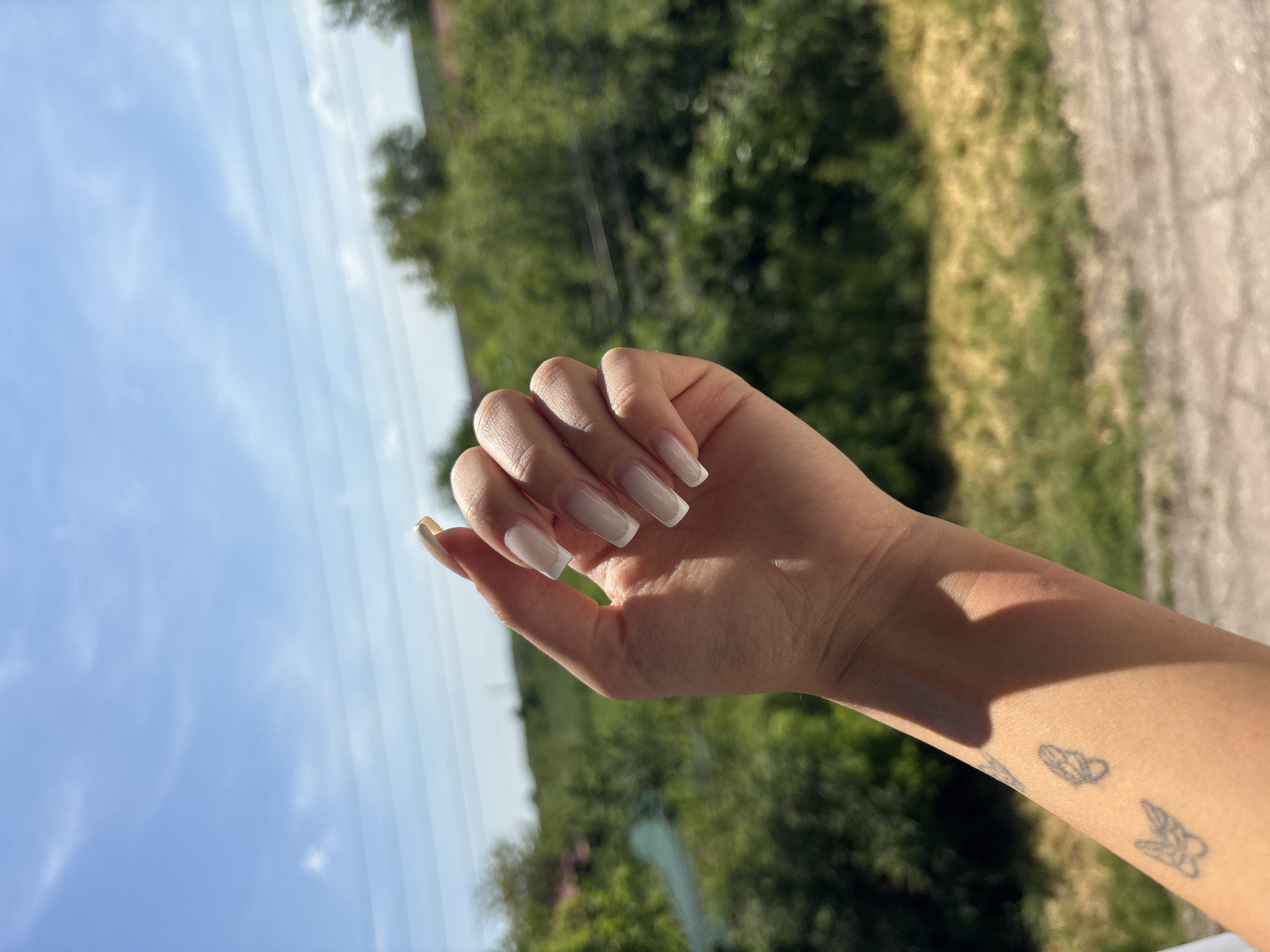 The image shows a hand with long, square-shaped white nails. The wrist of the hand is visible, bearing three small, simple tattoos: a feather, a cloud, and a flower. The background consists of green foliage, suggesting trees or bushes, and a blue sky with faint white lines. The scene appears to be outdoors, possibly in Medkovets, Bulgaria, as specified. There are no discernible actions or interactions depicted.