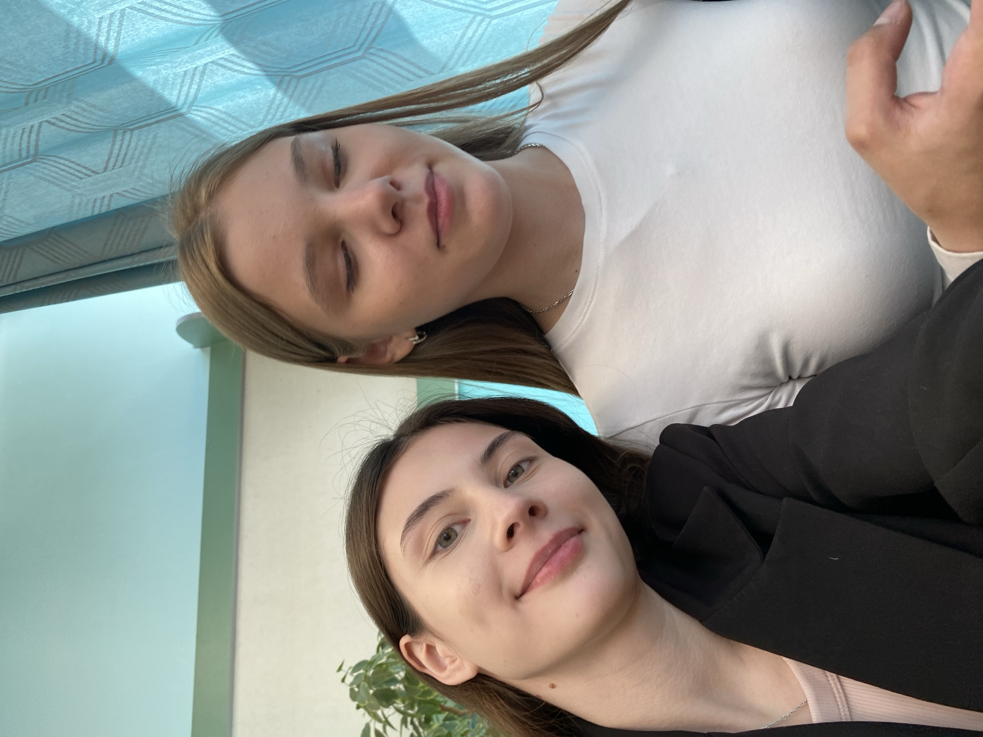 Two young women pose close together for a selfie. The woman in the foreground has dark brown hair, fair skin, and is wearing a black blazer over a light-colored top. She gazes toward the camera with a slight smile. The woman behind her has blonde hair, fair skin, and is wearing a white t-shirt. Her eyes are closed, and she also has a slight smile. The background includes a pale blue and white patterned curtain and a light green wall. The location is identified as Votkinsk, Russia. A green leafy plant can also be seen in the lower left corner.