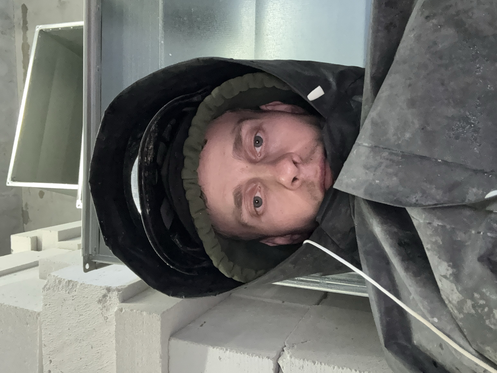 The image shows a close-up, selfie-style portrait of a man wearing protective gear, including a hood and possibly a helmet, that obscures much of his face and body. He appears to be in an industrial or construction environment. To the left of the man, there are stacked light-colored blocks, possibly concrete or cinder blocks, some of which are rough-textured. To the right and behind him, there is a large, metallic ductwork system, appearing to be part of ventilation or HVAC infrastructure, with a rectangular opening visible in the upper section. The lighting is somewhat harsh and utilitarian, suggesting an indoor setting with artificial light. There are no overt indications of the specific location within Moscow, Russia, beyond the general context of an urban industrial or construction site.