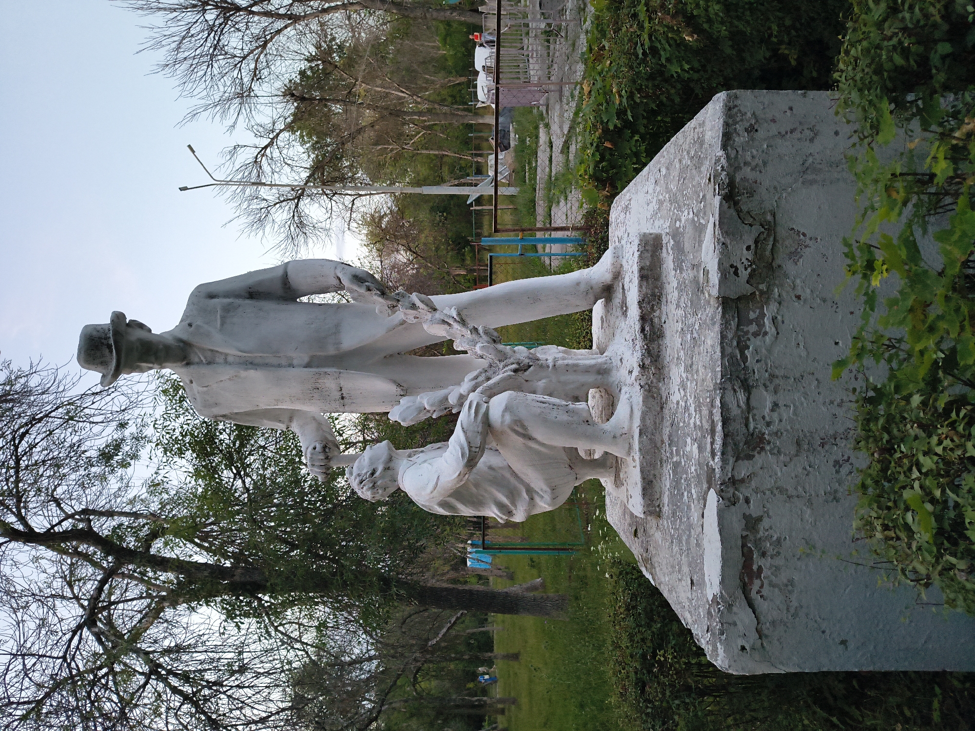 A white concrete statue depicts two figures in an outdoor park setting. The taller figure, standing upright and wearing a hat and suit, is looking down. A shorter figure, kneeling, looks up towards the standing figure and is holding a branch or wreath. The statue is placed on a weathered concrete pedestal surrounded by greenery. In the background, trees are visible, some with bare branches, and a lamppost stands in the middle ground. A fence with a gate can be seen behind the statue. The overall scene suggests an overcast day in a public park. This statue is located in the city of Mikhaylovka, Russia.