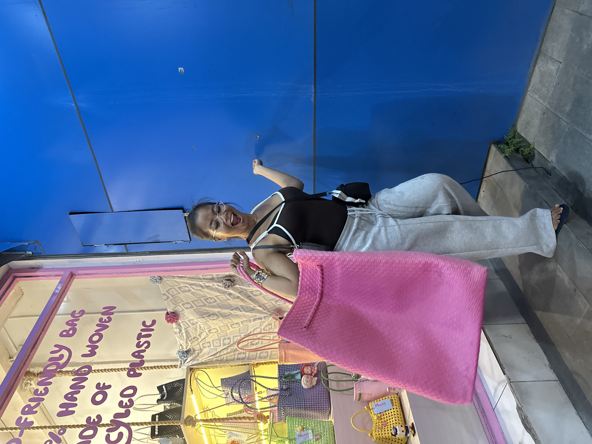 The image shows a woman in Ubud District, Indonesia, posing in front of a shop selling bags. She is wearing glasses, a black top with white straps, and light-colored sweatpants. She holds a large pink woven bag. The shop window displays various handmade, woven bags in different colors. The sign on the window reads "Eco-Friendly Bag Hand-Woven of Styled Plastic". The backdrop is a blue wall.