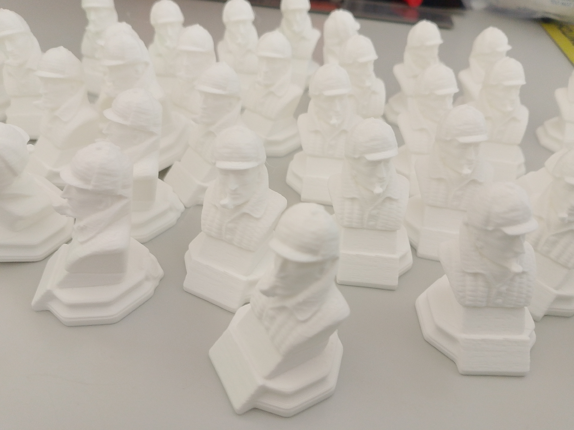 The image shows a collection of 3D-printed, white busts of Mikhail Kalashnikov, the designer of the AK-47 rifle. The busts are situated on a light grey surface. The setting is not explicitly identifiable as Izhevsk, Russia, based on visual information alone. The busts are depicted in detail, with each wearing a cap and a jacket. There are no people visible in the image besides the subjects of the busts.