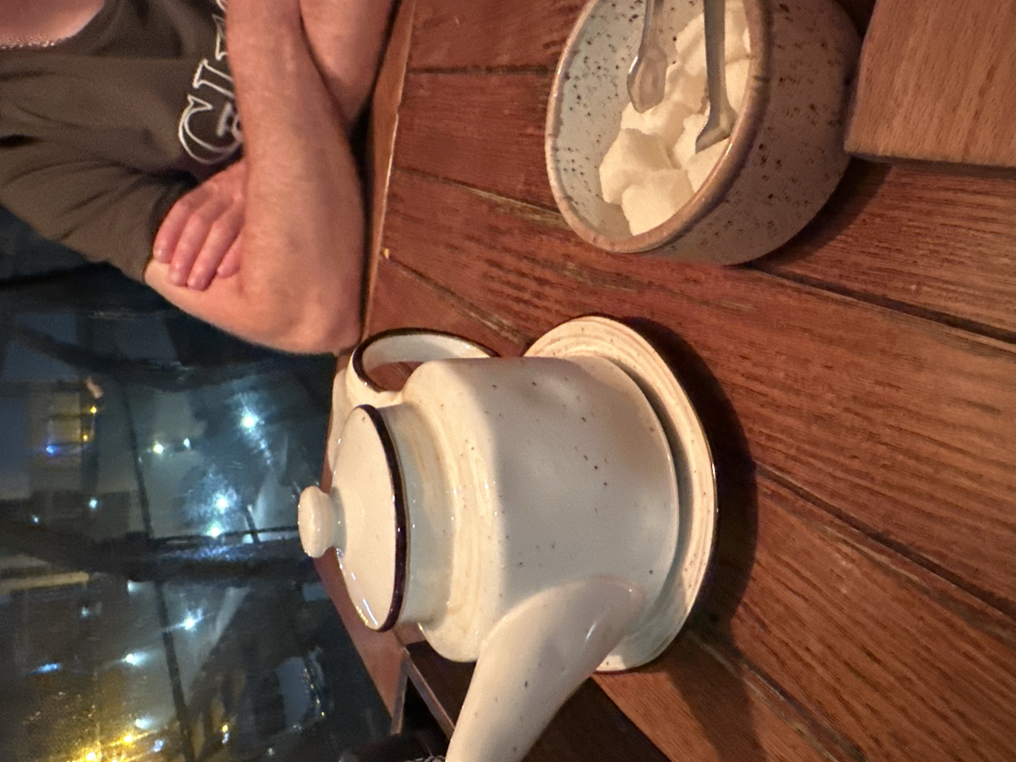 A close-up shot shows a white ceramic teapot with a dark rim and speckled detailing, resting on a matching saucer on a wooden table. To the right of the teapot, a small, speckled ceramic bowl contains white sugar cubes and a pair of silver tongs. In the background, a person wearing a dark green t-shirt with a white logo is visible, with their arms crossed. The scene appears to be set indoors, possibly in a restaurant or cafe, with a window in the background showing a blurry view of city lights at night, suggesting a location in Sochi, Russia.