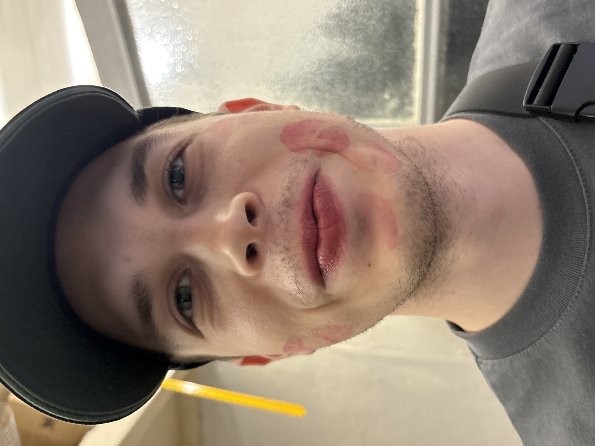 A young adult male is depicted in a close-up, head-and-shoulders portrait shot within an indoor setting in Saint Petersburg, Russia. He wears a dark baseball cap and a grey, short-sleeved t-shirt. Distinct red lipstick marks are visible on both cheeks and around his lips, with a particularly prominent mark on his right cheek (viewer's left). Light stubble covers his jawline and upper lip area. He looks directly at the camera with a subtle smirk or smile. A black strap with a buckle, possibly from a bag, is visible over his right shoulder. The background on the left features a light-colored wall with hints of wooden structural elements and an out-of-focus yellow object. The background on the right shows a textured, light-colored surface, possibly a window with condensation or patterned glass, illuminated from behind.